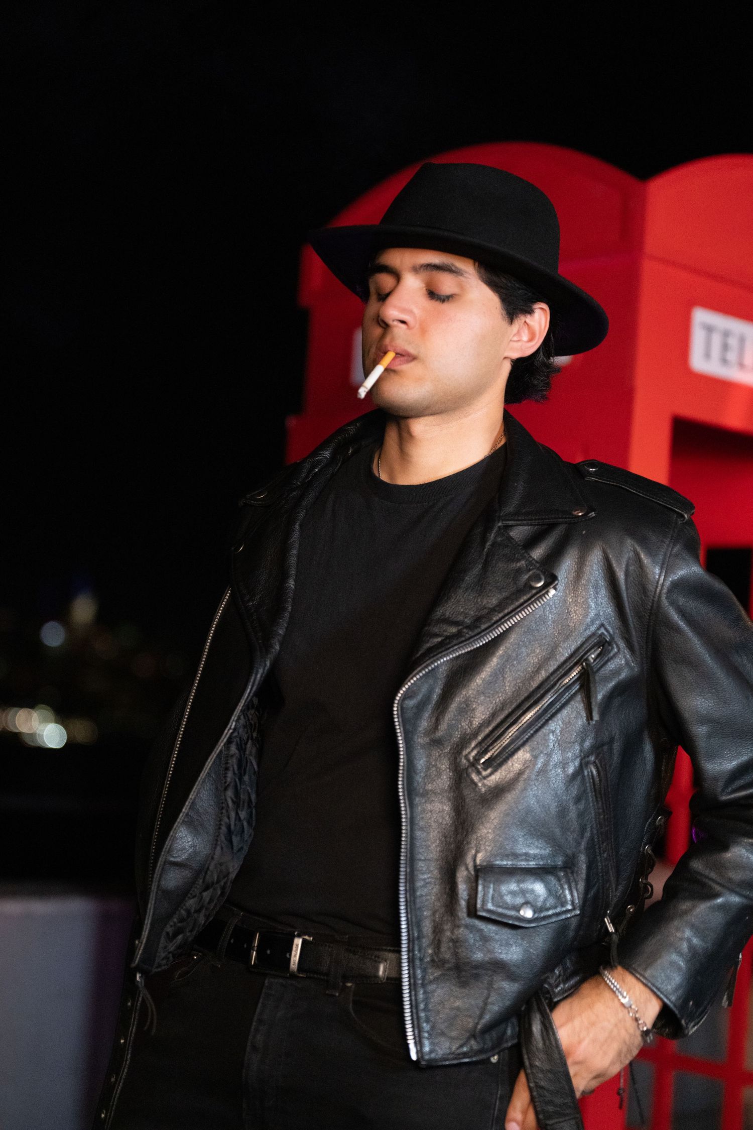 Person in black leather jacket and hat strikes a dramatic pose against red background.