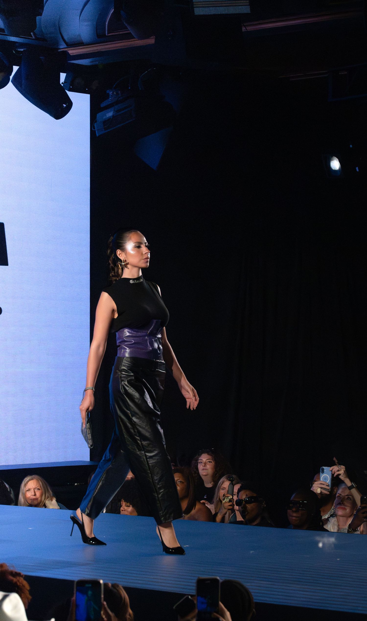 Model in black leather dress walks runway under dramatic lighting.