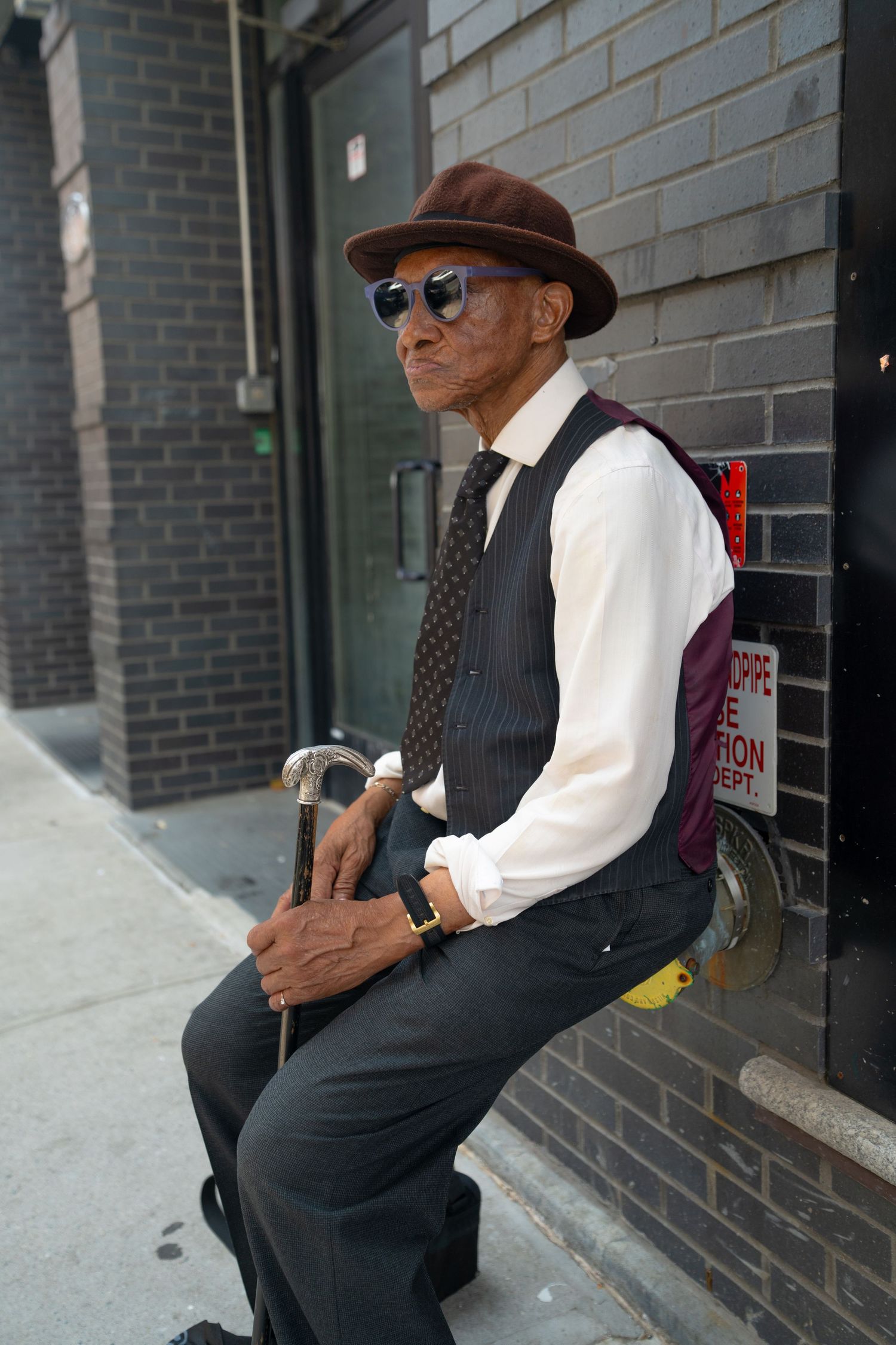 Person in brown hat and formal attire sitting on steps against a brick wall.