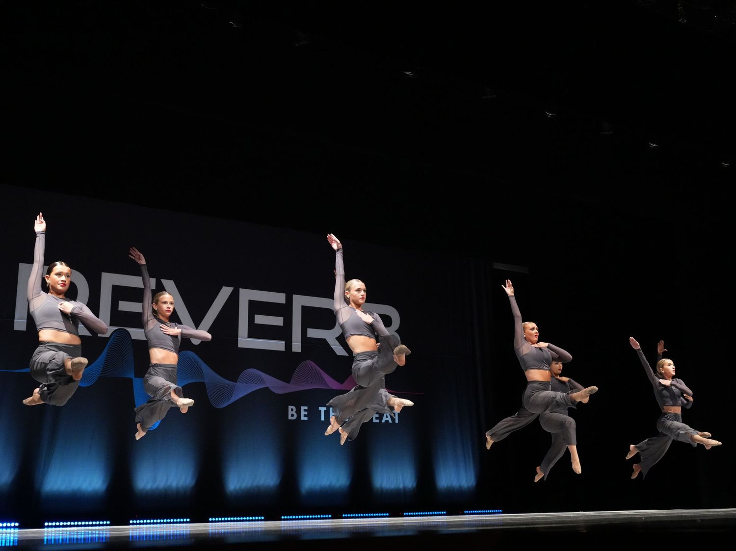Five dancers leap mid-air on stage in gray outfits before a blue Reverb Be The Beat backdrop.