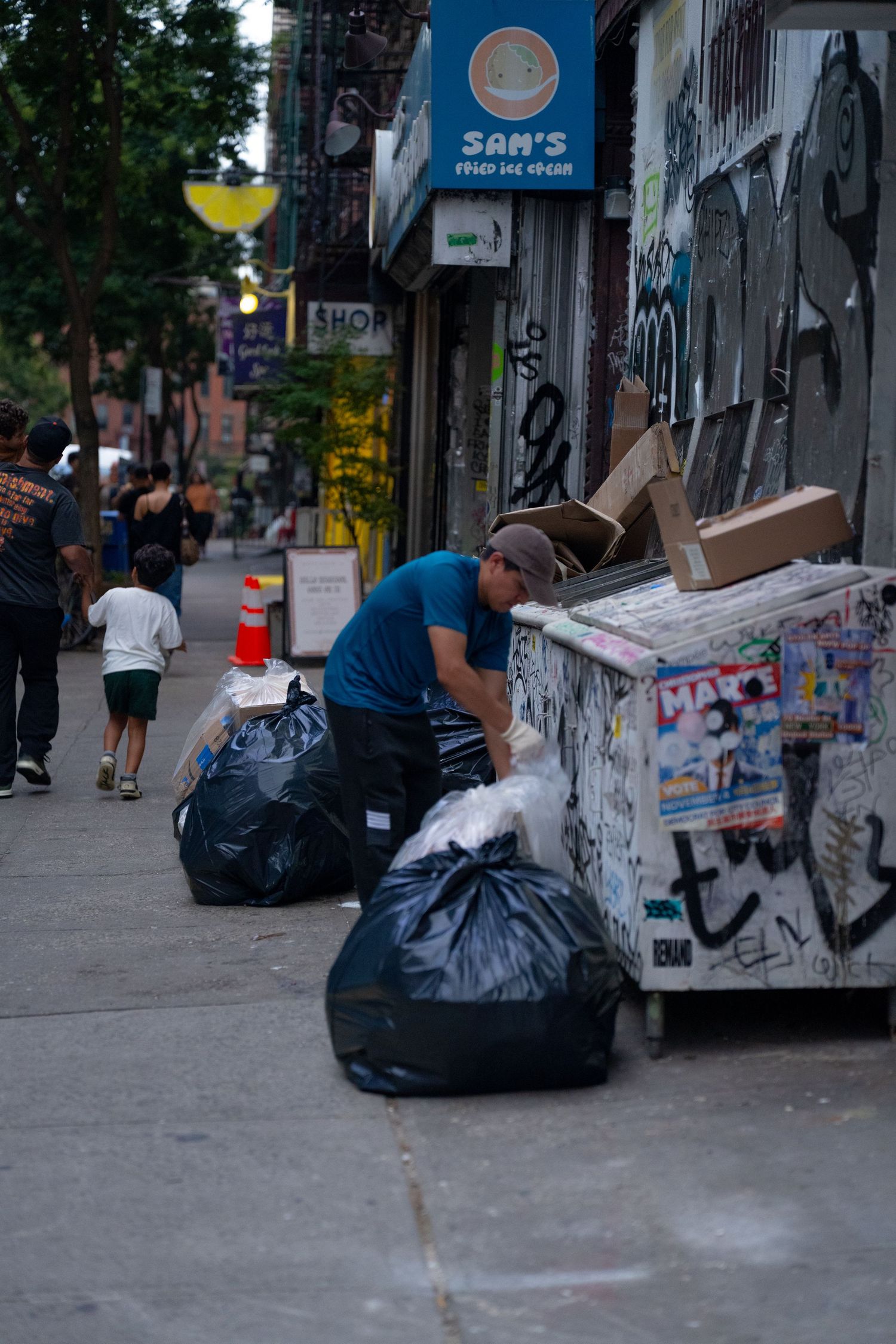 Black garbage bags line a city sidewalk against graffiti-covered walls near Sam's store.