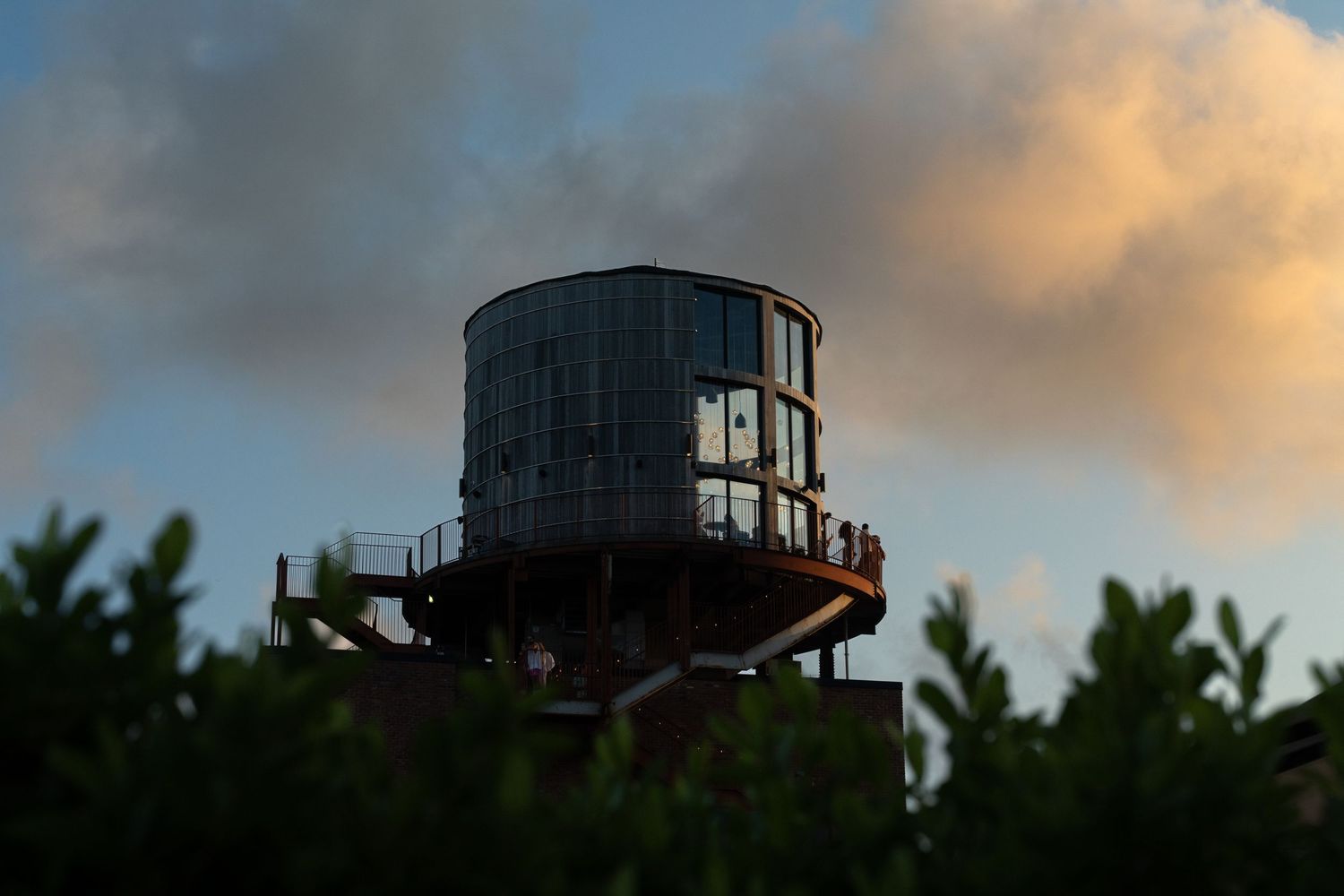 Rustic water tower with observation deck surrounded by lush green trees against a dramatic sunset sky.