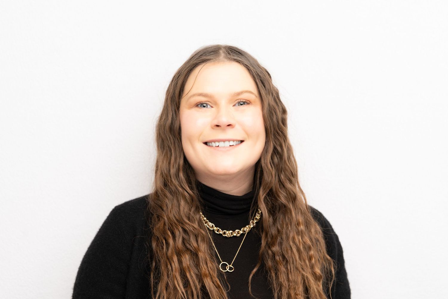 Professional headshot of a person with long brown wavy hair wearing a black sweater and gold necklaces against white background.