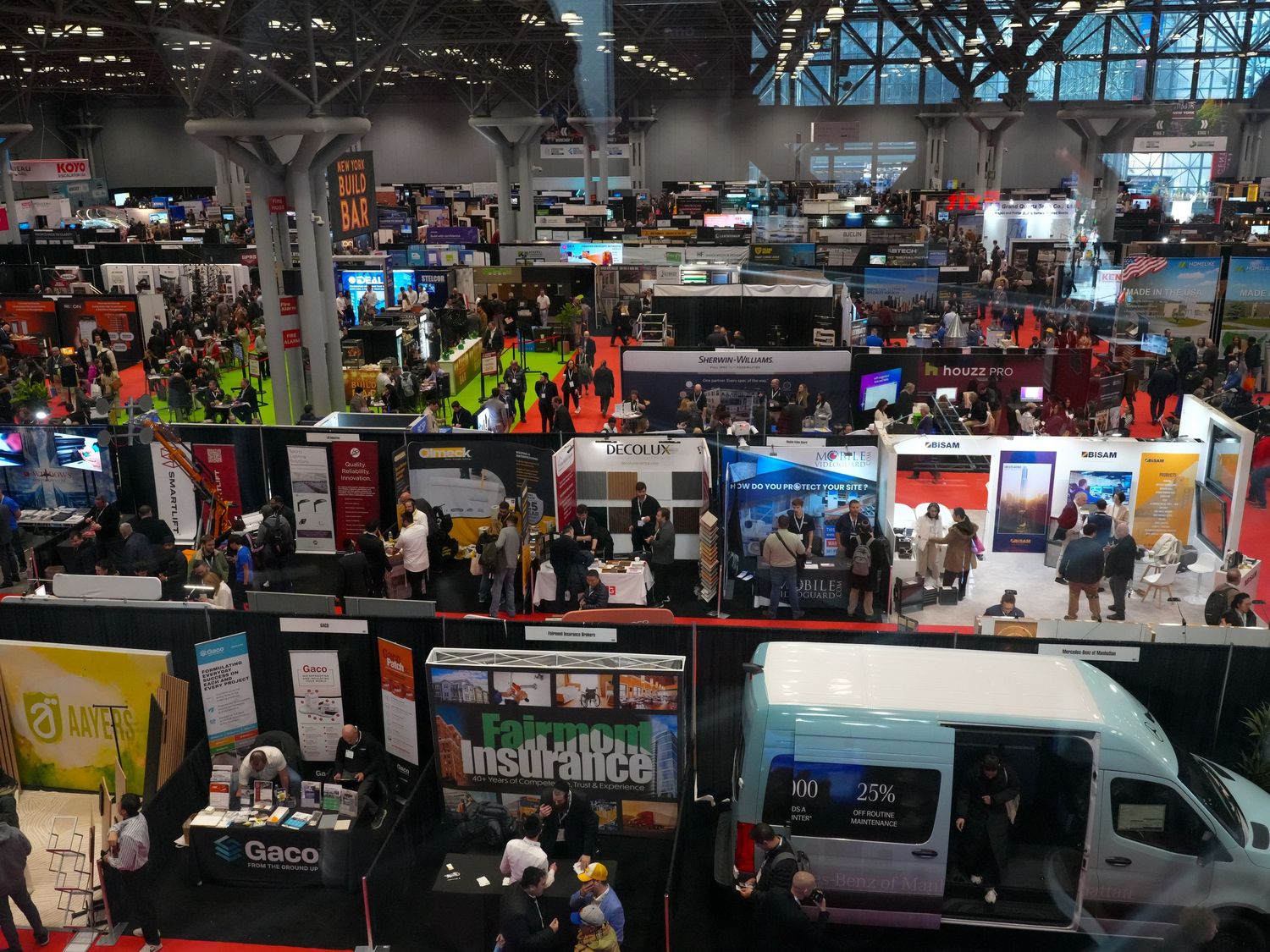 Aerial view of a busy trade show floor with vendor booths, crowds, and a white van at a large convention center.