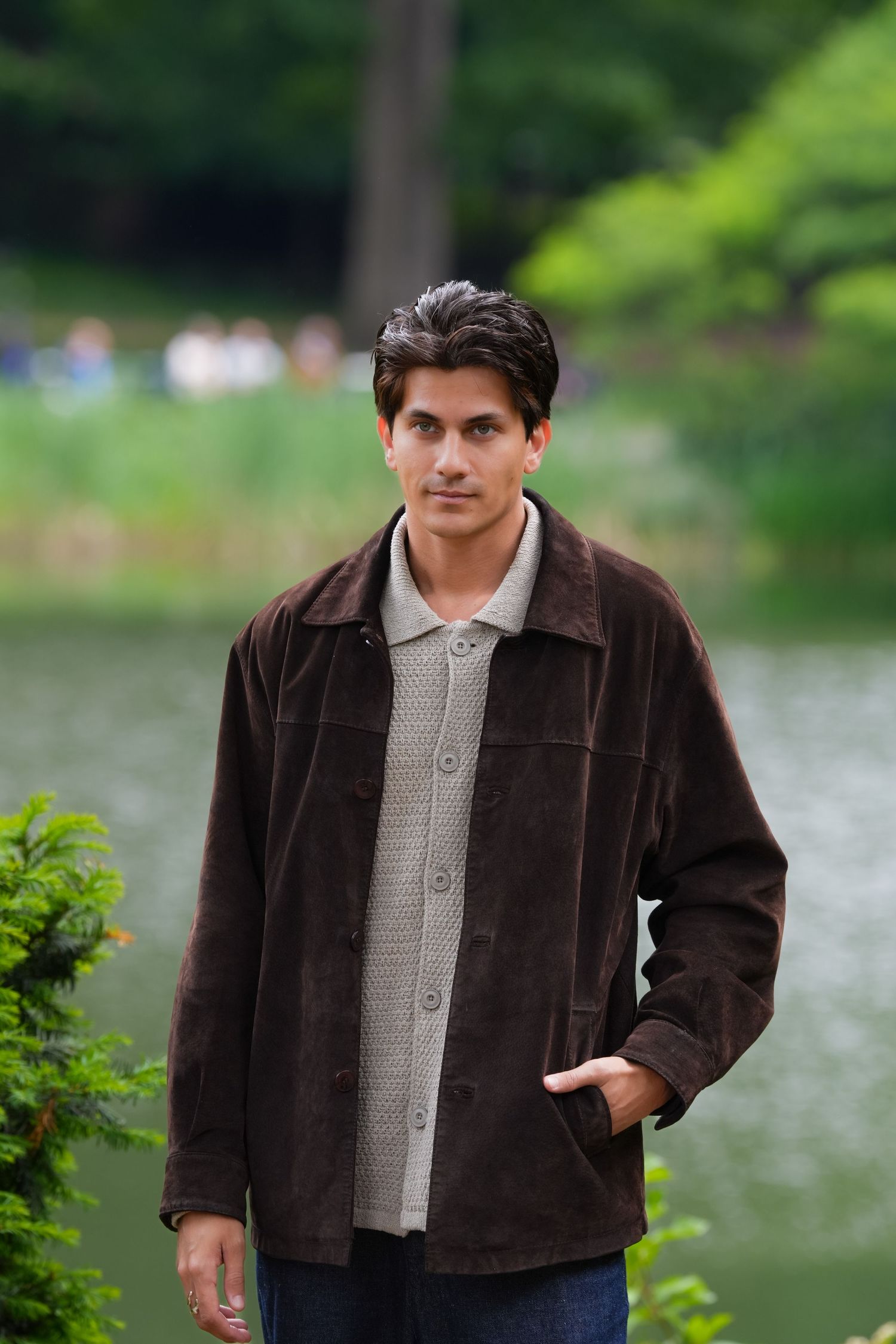 Young man in brown suede jacket and gray sweater standing by a serene park lake with lush green trees.