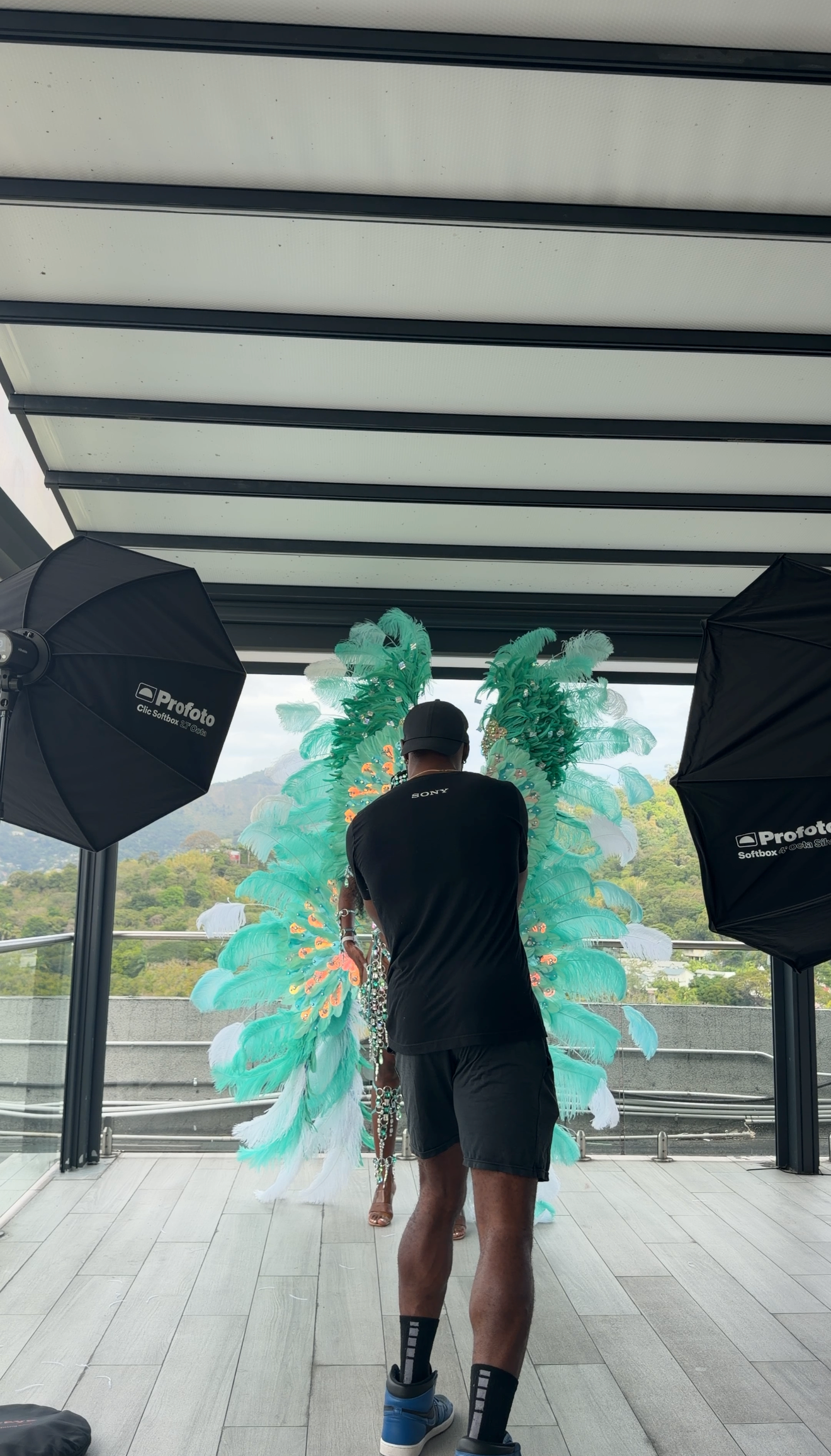 Photographer with Profoto lights shooting model wearing large teal feathered carnival wings on rooftop terrace.