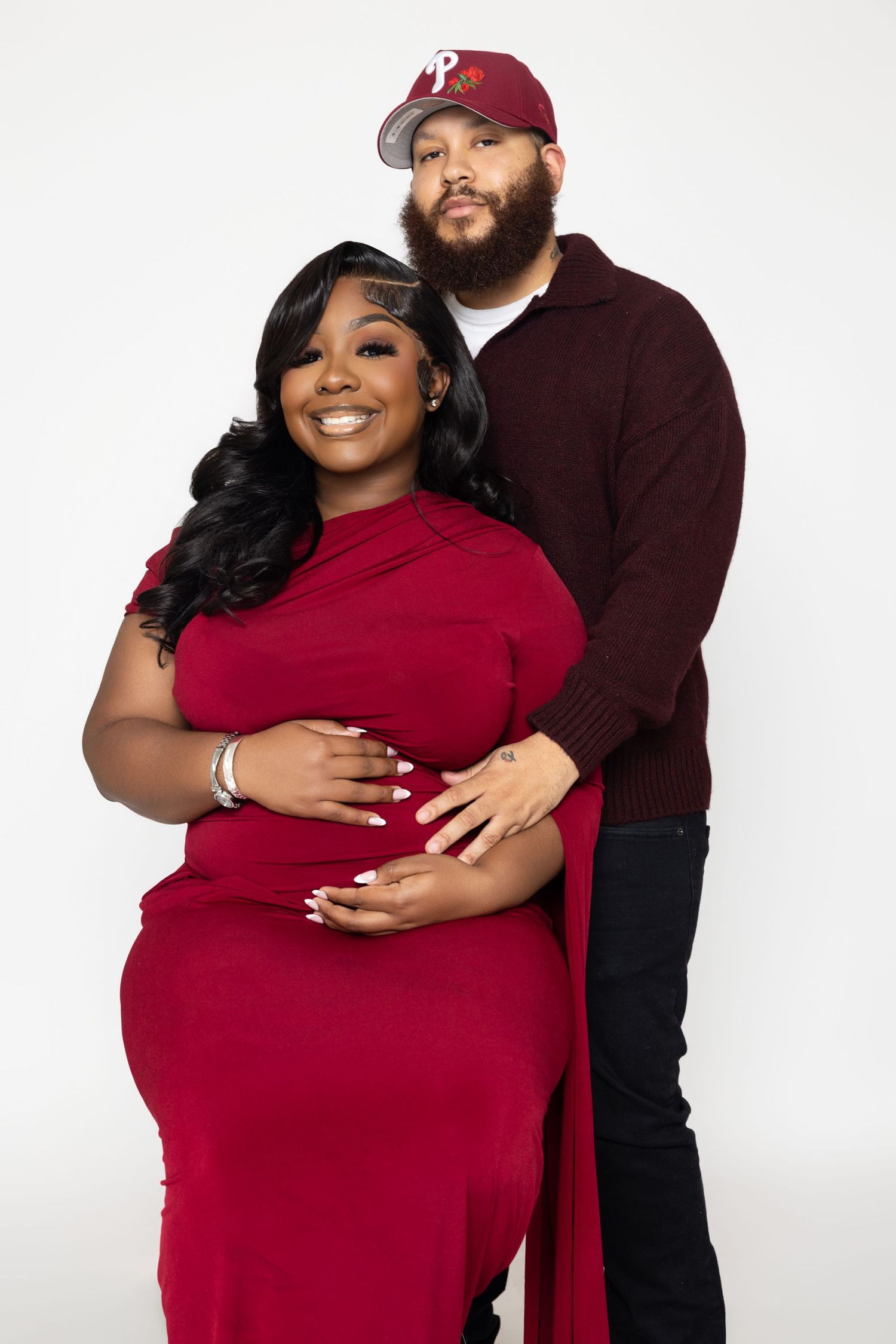 A couple poses together against a white backdrop in coordinating burgundy outfits for a romantic photoshoot.