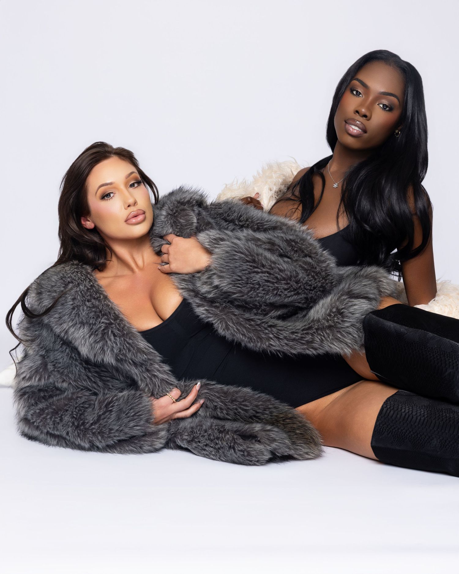 Two models in black outfits pose with grey fur coat on white background for fashion shoot.