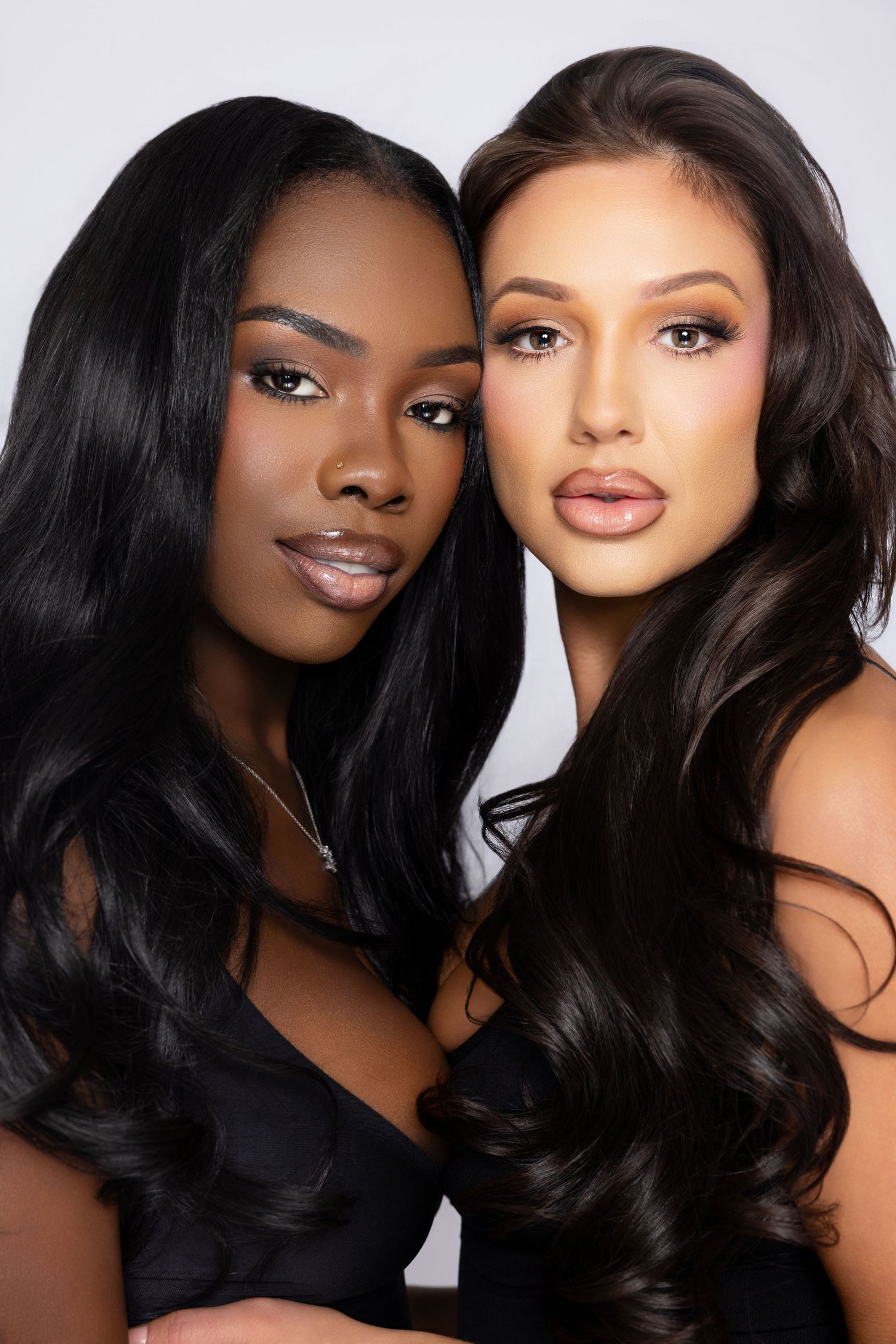 Two models with long dark hair pose together wearing black tops against a light background for a beauty photoshoot.