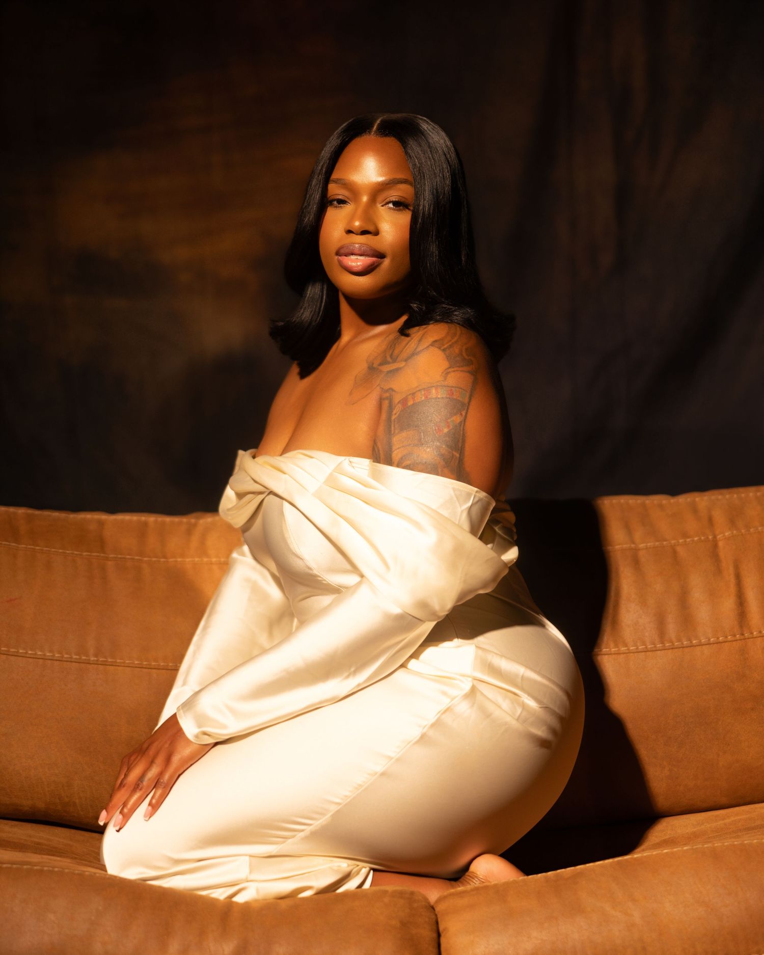 Woman in elegant off-shoulder satin outfit seated on a brown couch against a dark backdrop.