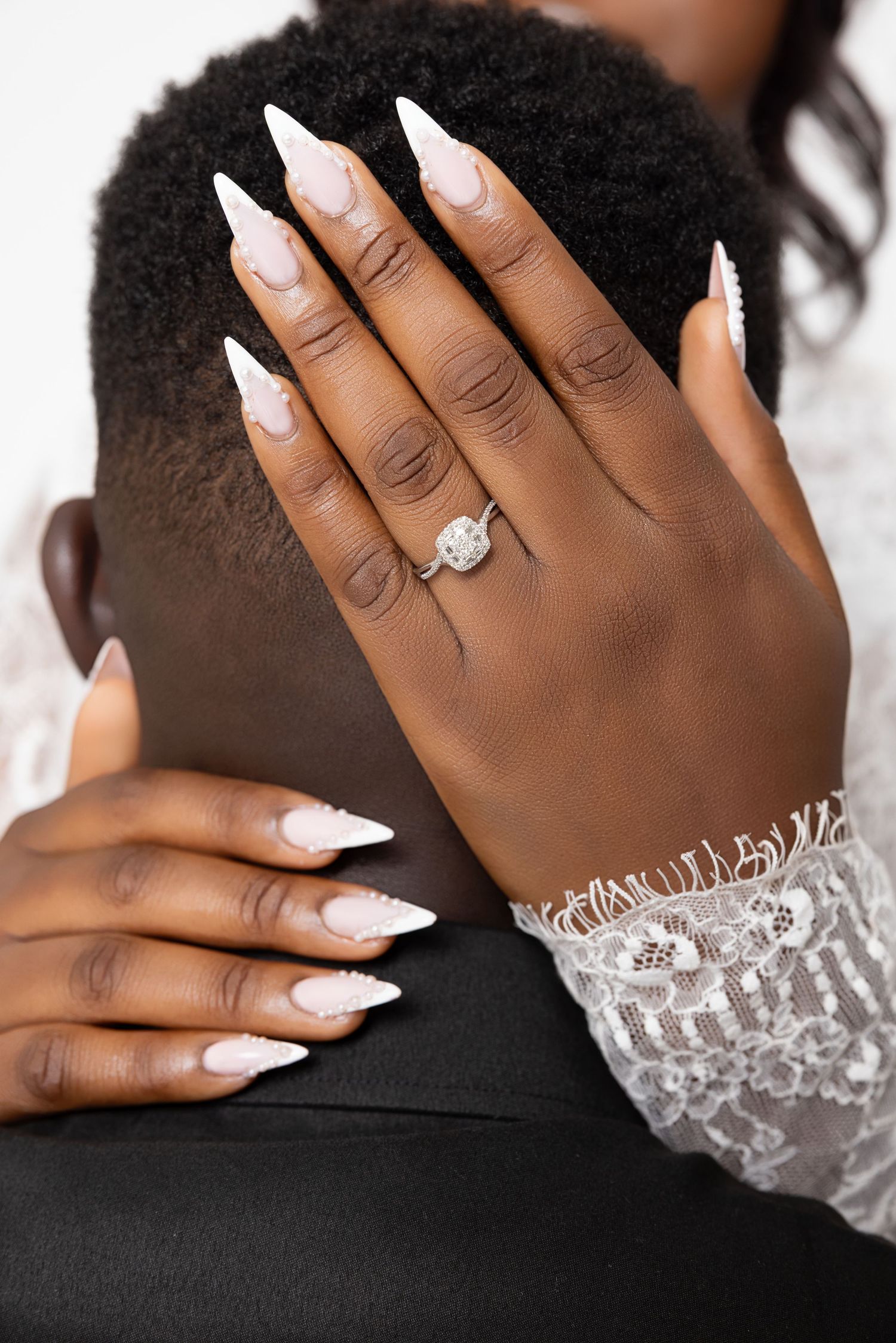 Close-up of hands with white manicured nails and a sparkling engagement ring against black fabric.