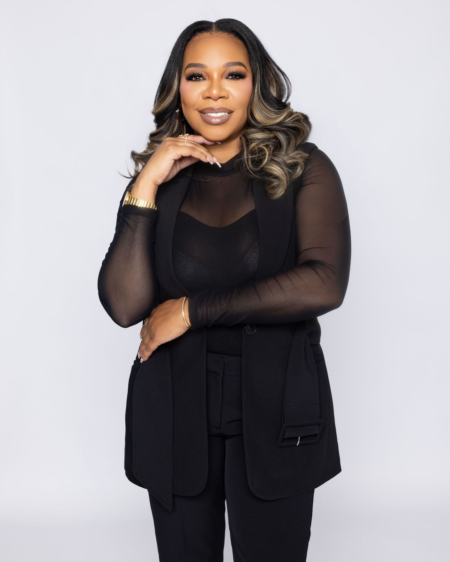 A professional woman in an all-black outfit poses confidently with her hand on her chin against a light background.