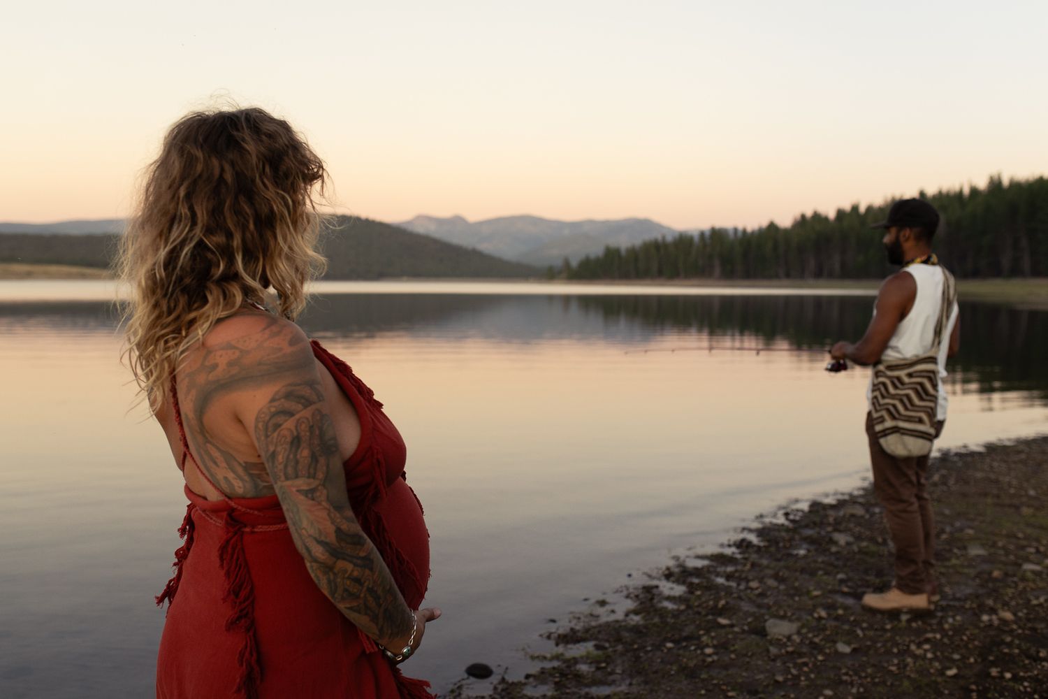 Couple at a calm mountain lake at golden hour, woman in a red dress watching her partner cast a fishing line, relaxed engagement session