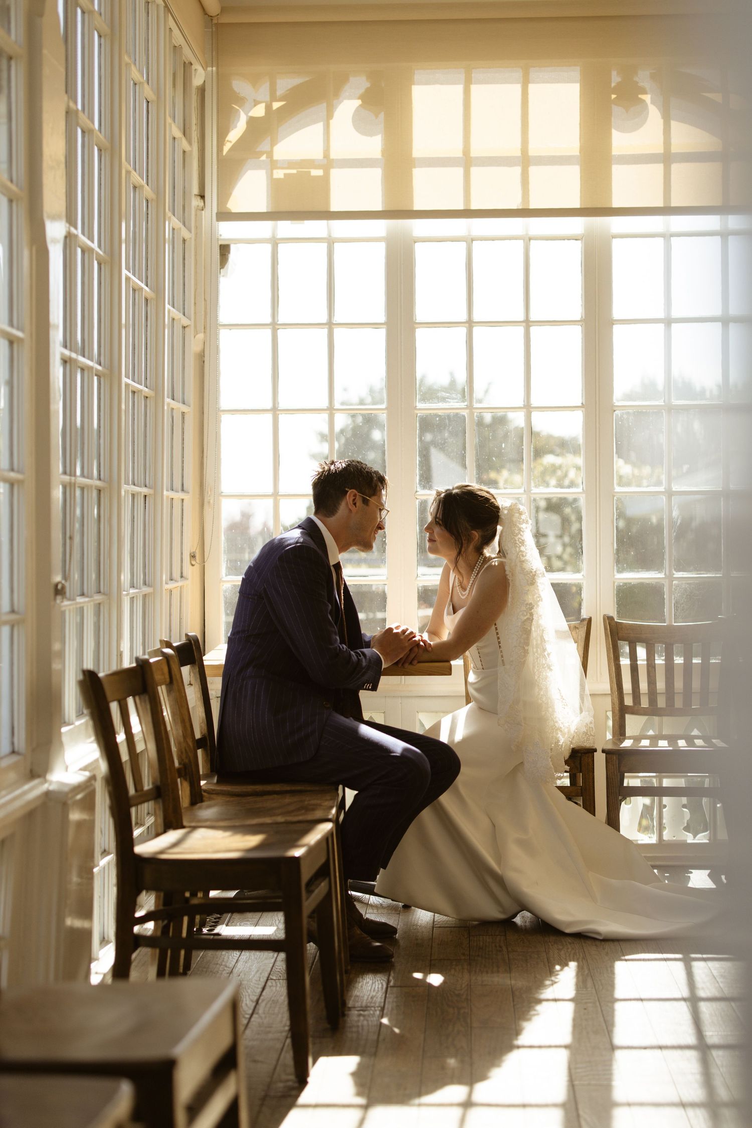 Couple sitting together by a tall sunlit window in a rustic room, warm and intimate wedding portrait