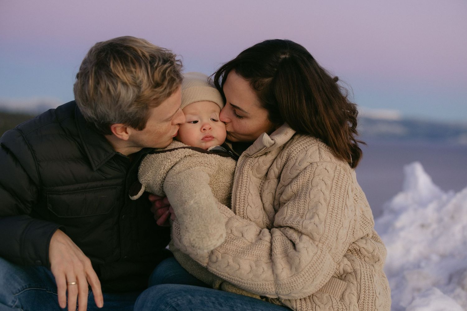 Couple kissing their baby's cheeks at sunset above a snowy mountain lake, joyful Lake Tahoe family session