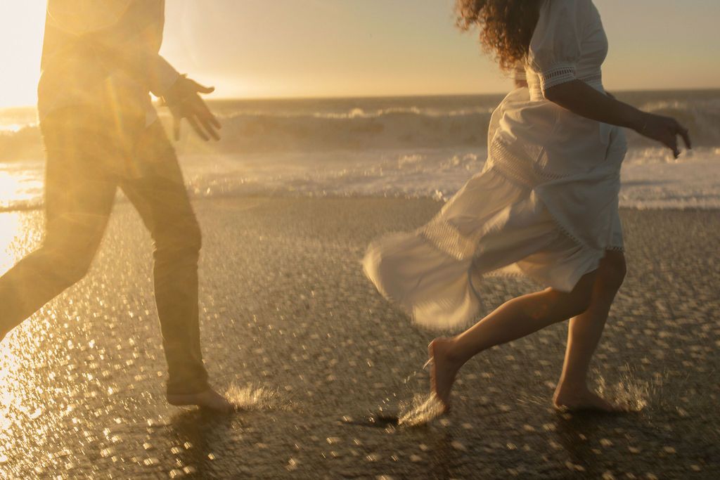 Couple playing on the wet sand as waves and sun shimmer behind them, joyful coastal couples session