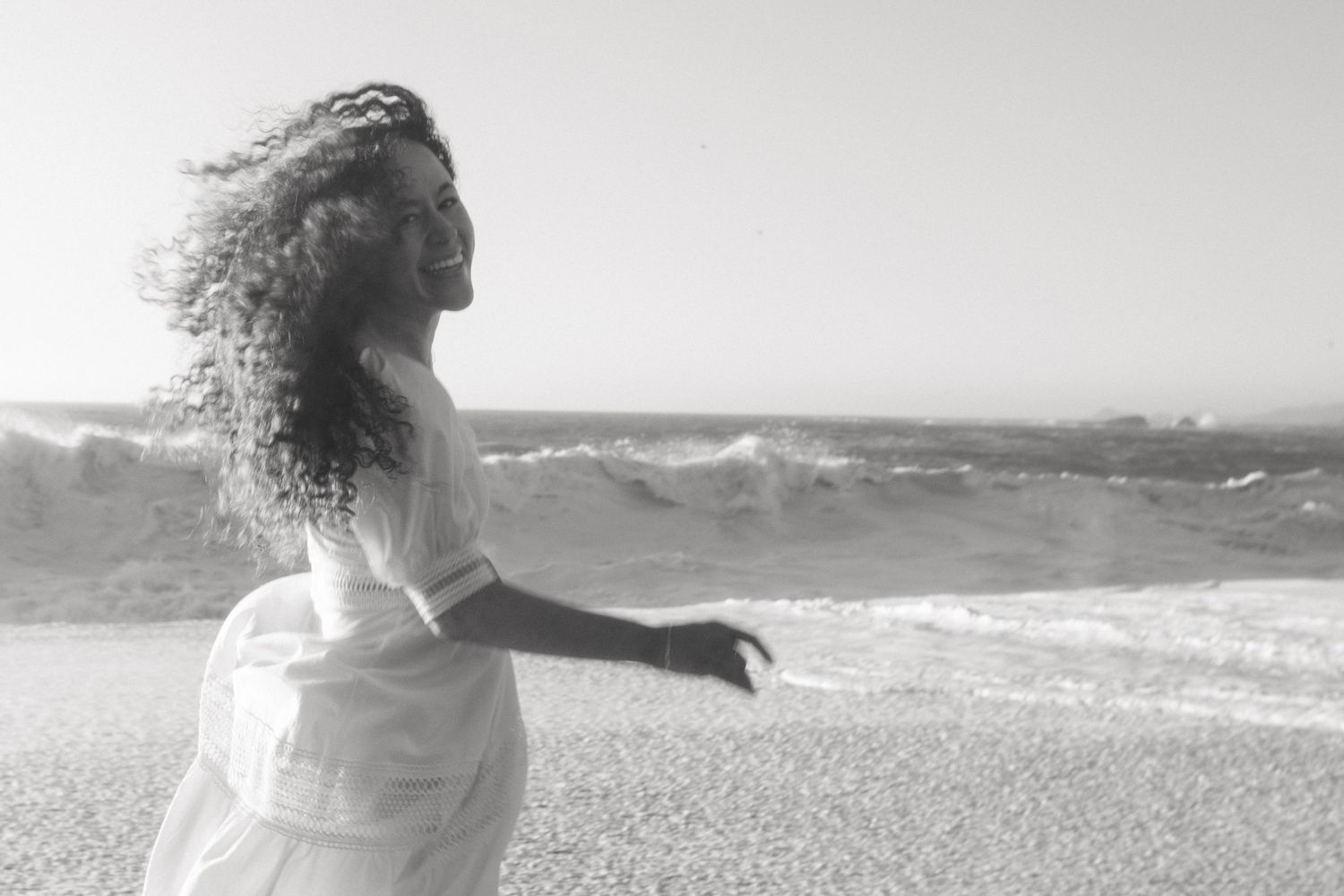Woman twirling joyfully in white dress on beach with ocean waves during engagement session in Gualala California Mendocino CA by Sammy Aucel