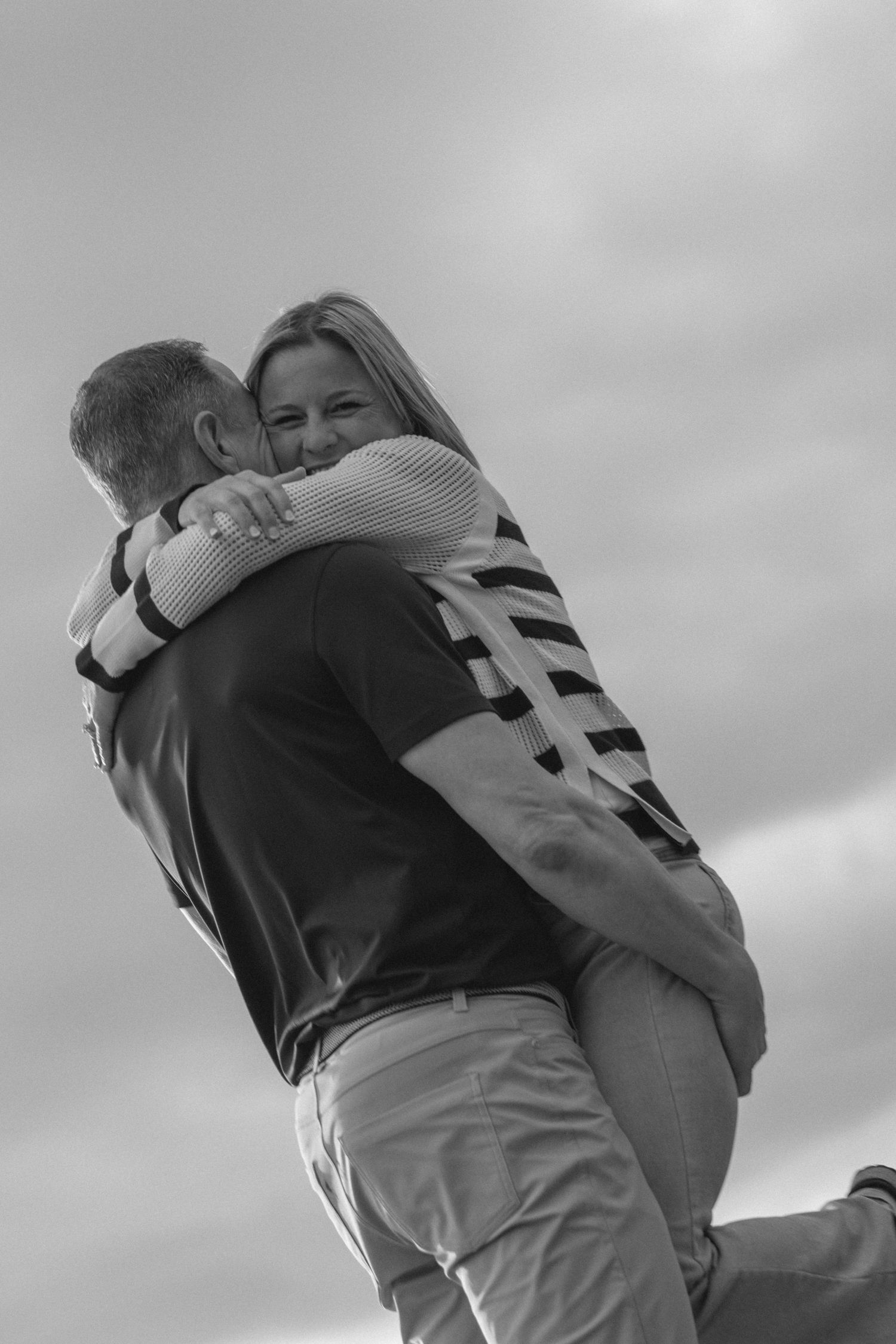 Black-and-white photo of a man lifting his partner in a joyful embrace against a cloudy sky, candid couples session