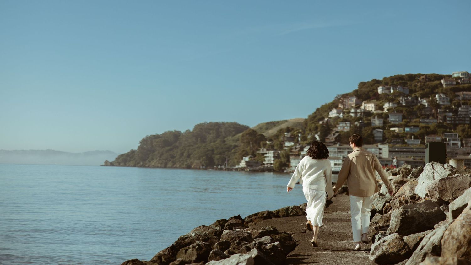 Couple walking a coastal path above the bay with a hillside town beyond, scenic San Francisco engagement session