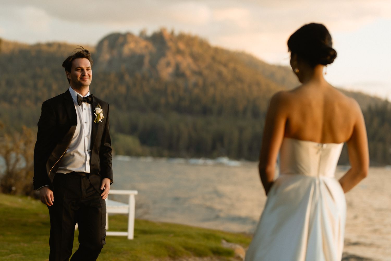 Bride in an open-back gown facing her groom beside an alpine lake at golden hour, romantic mountain wedding portrait