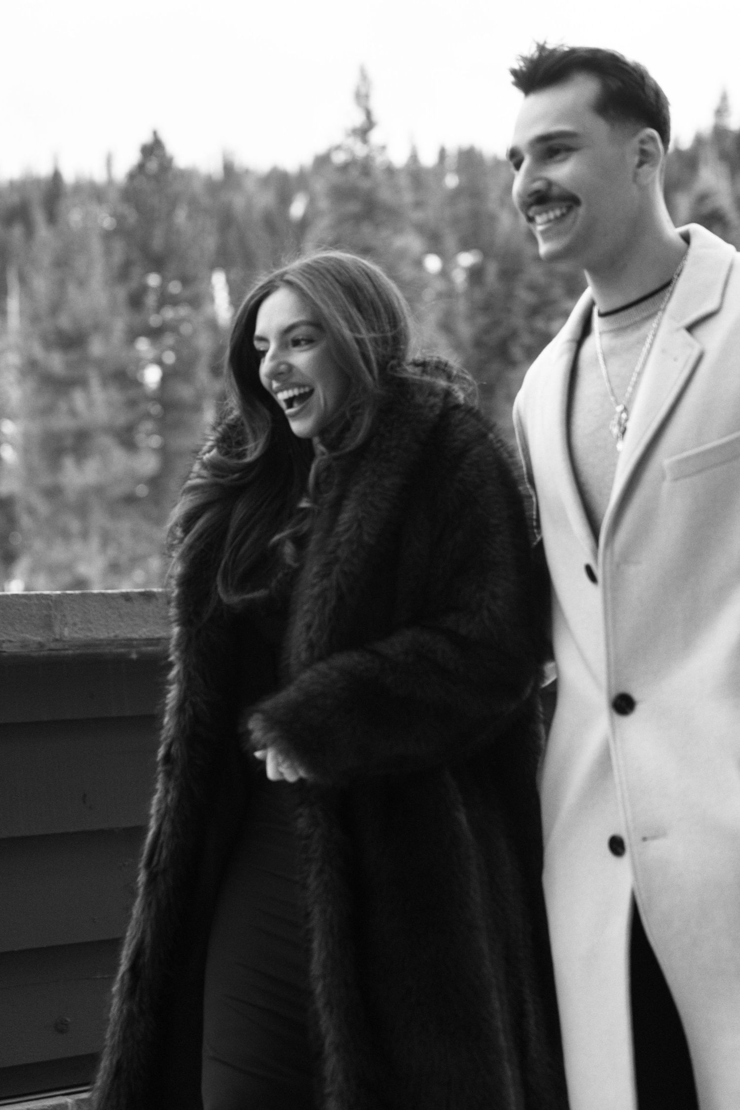 Black-and-white photo of a stylish couple in winter coats laughing together among snowy pines, chic cold-weather engagement session