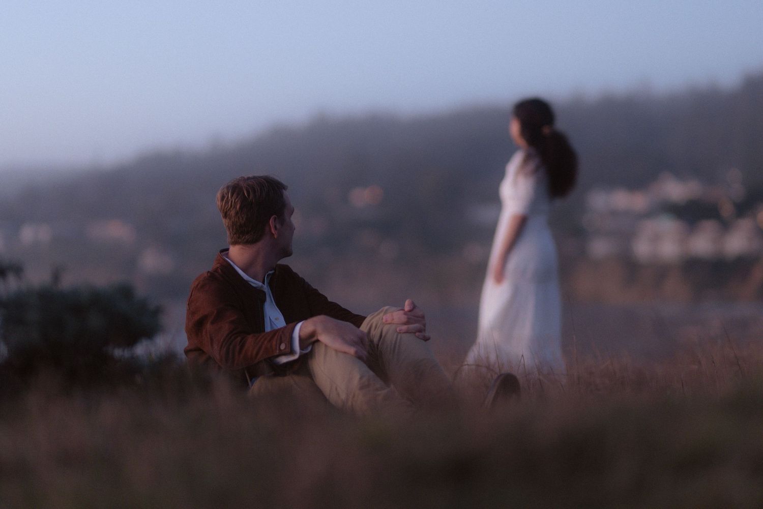 Couple sharing a glance on a coastal bluff at dusk with the bay behind them, romantic Northern California engagement session