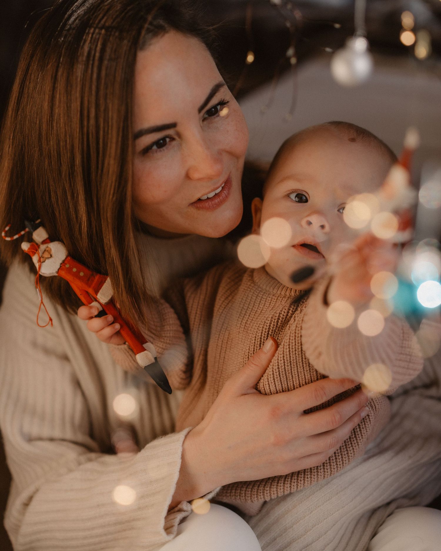 Mother cradling her smiling baby amid warm twinkling bokeh lights, cozy lifestyle family session
