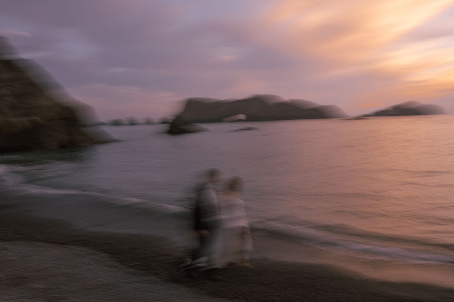 Couple as dreamy motion-blurred figures on a beach at sunset by coastal sea stacks, Harbor House Mendocino wedding