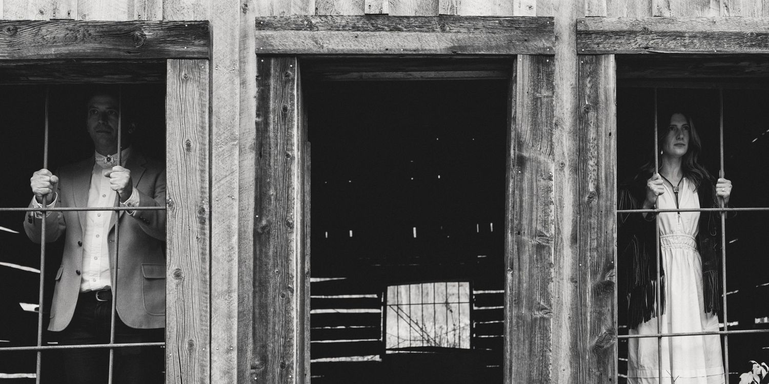 Black-and-white photo of a couple framed in the broken windows of a ghost-town building, moody desert engagement session