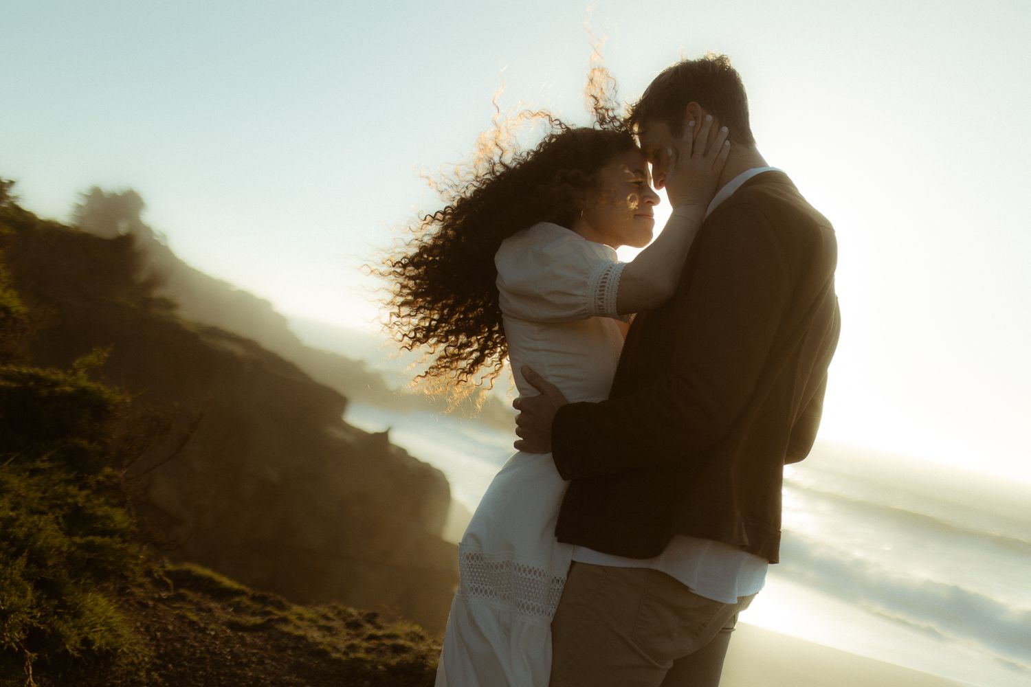 Couple resting forehead to forehead on a windswept coastal bluff at sunset, romantic elopement photography