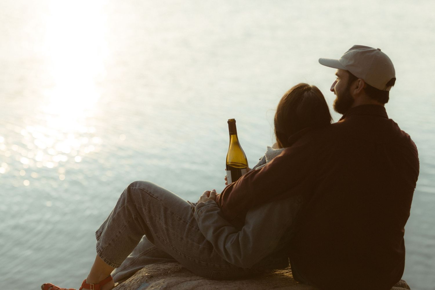 Couple cuddling on a lakeshore at golden hour with a bottle of wine, sun sparkling on the water, romantic engagement session
