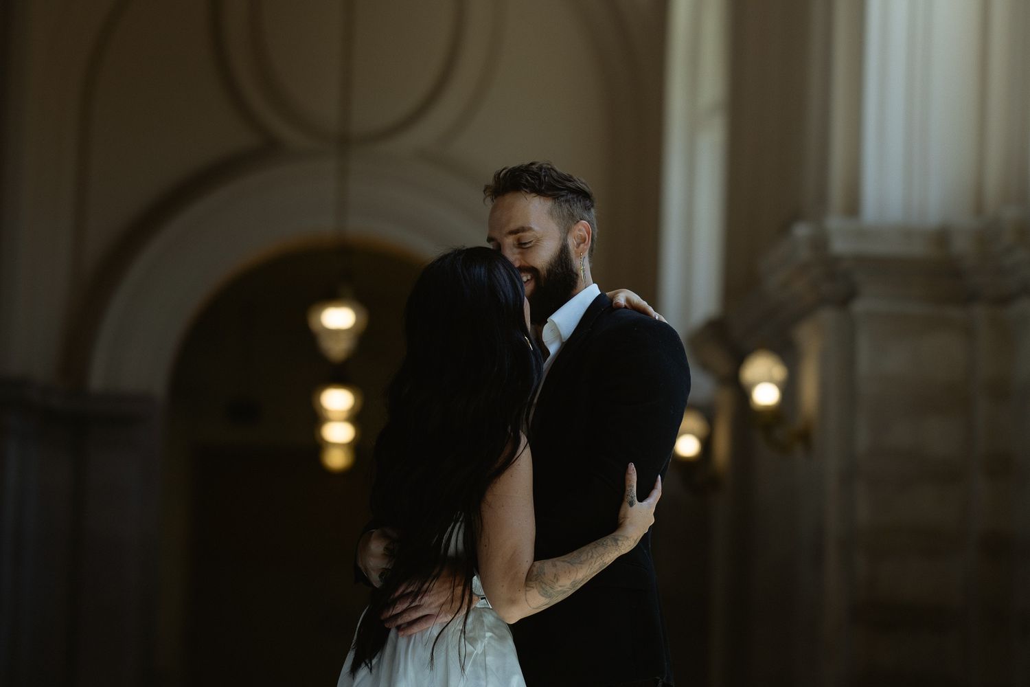 Couple embracing in a warmly lit arched hallway, bride in white and partner in a dark suit, intimate wedding portrait