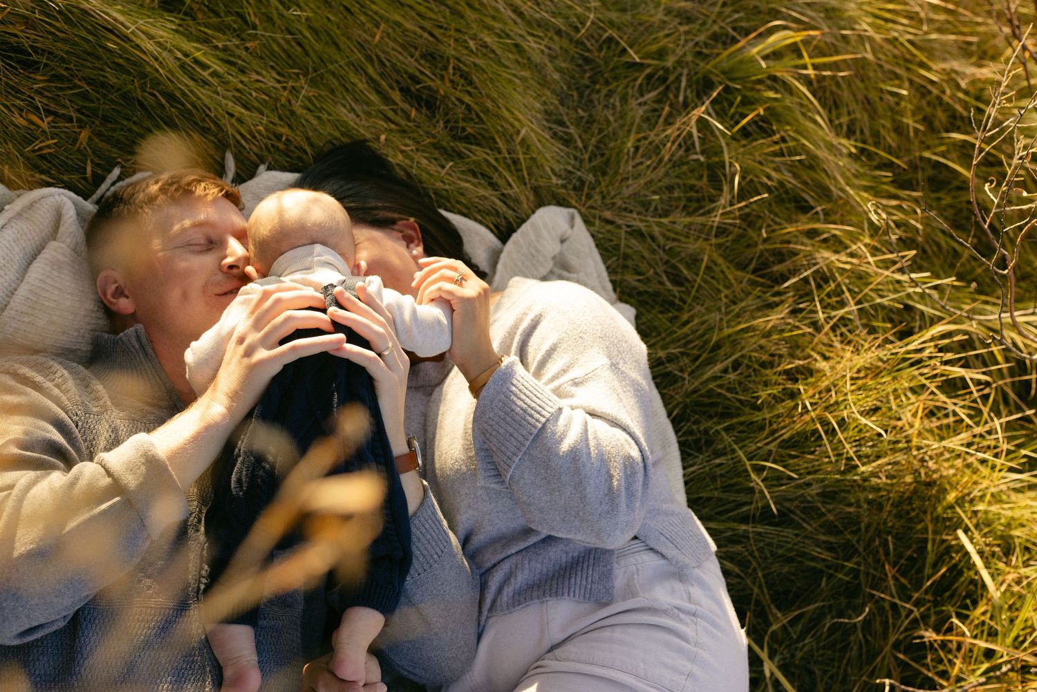 Parents lying in tall golden grass kissing their baby in the warm evening light, intimate outdoor family session
