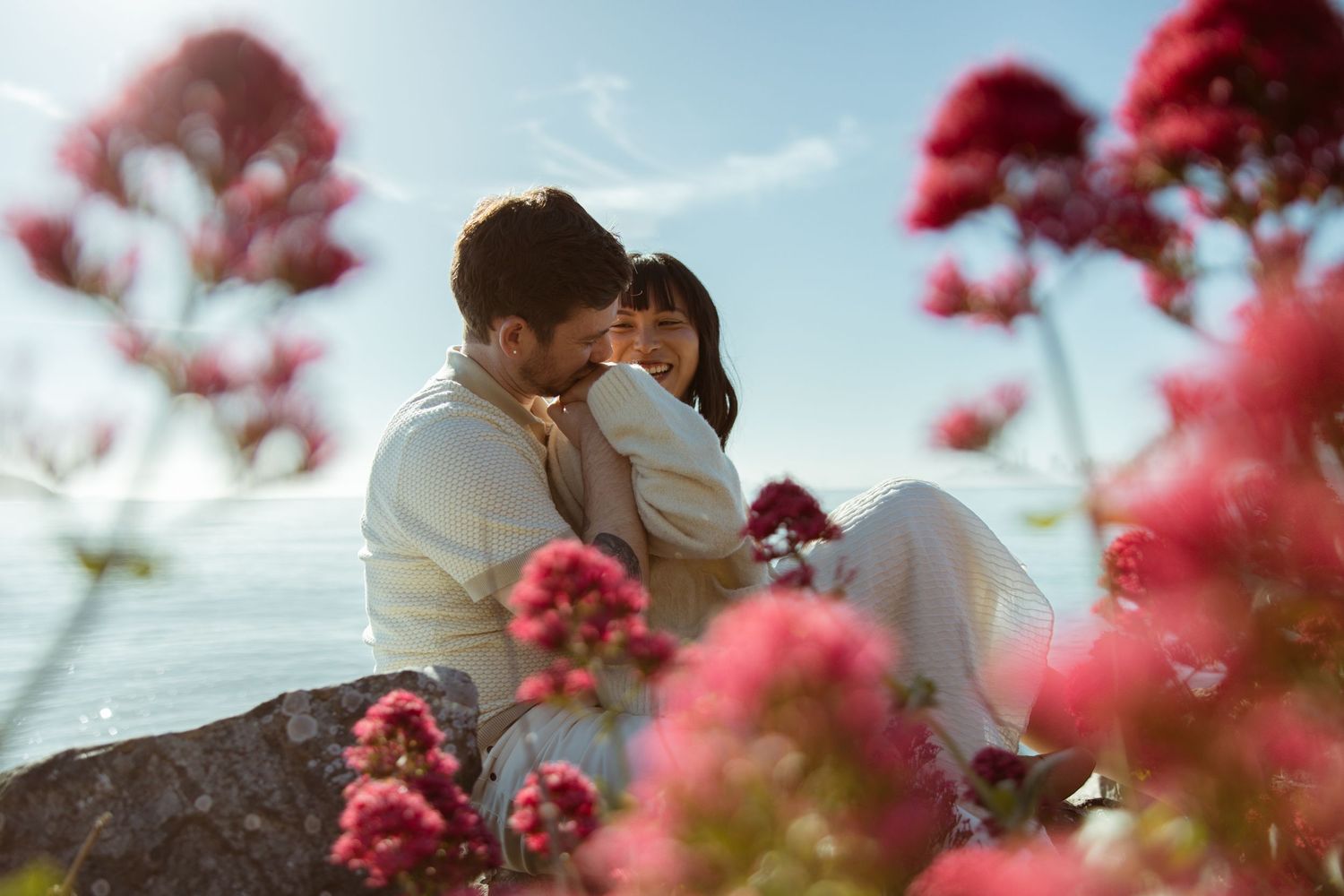 Couple embracing by the water framed through bright pink wildflowers, romantic coastal engagement session