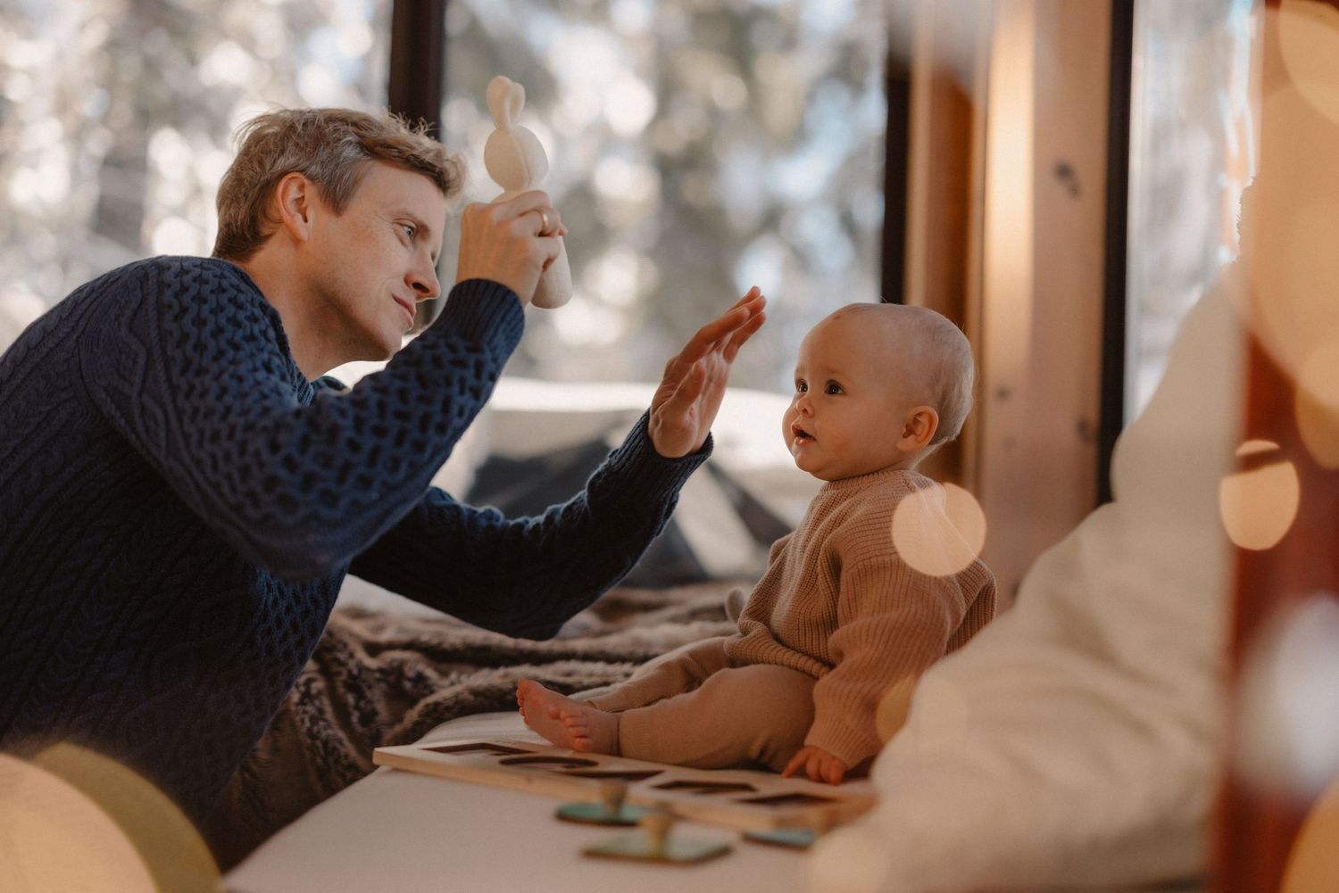 Father playing and high-fiving his baby inside a cozy cabin with snowy windows, joyful winter family session
