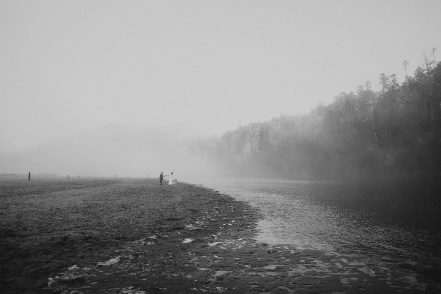 Black-and-white photo of a couple as tiny figures on a misty foggy beach at dawn, atmospheric coastal elopement