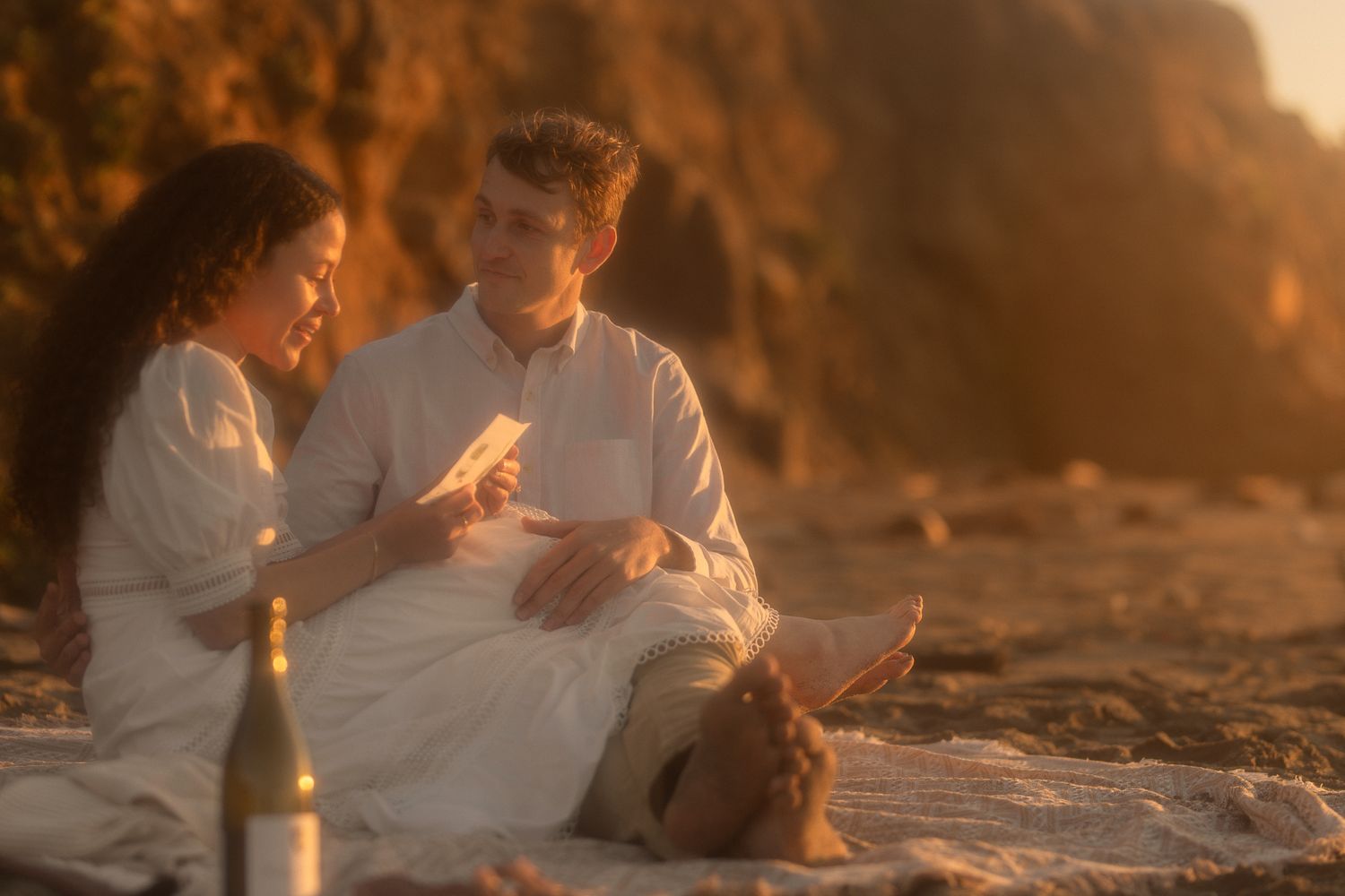 Couple sitting close together on the beach in warm golden light, romantic coastal engagement session