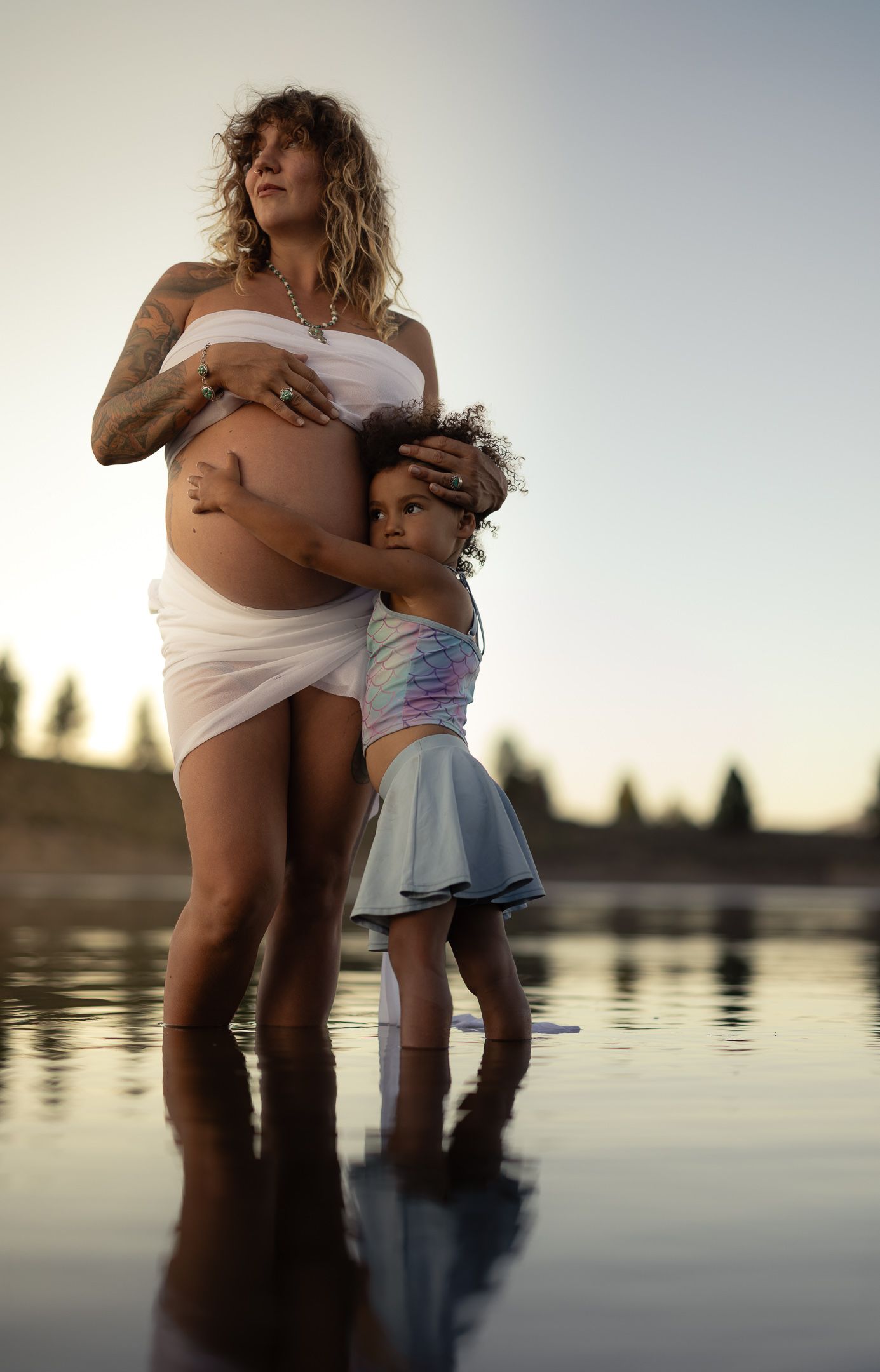 Expectant mother in white at a lakeside at sunset with her young child hugging her belly, glowing maternity session