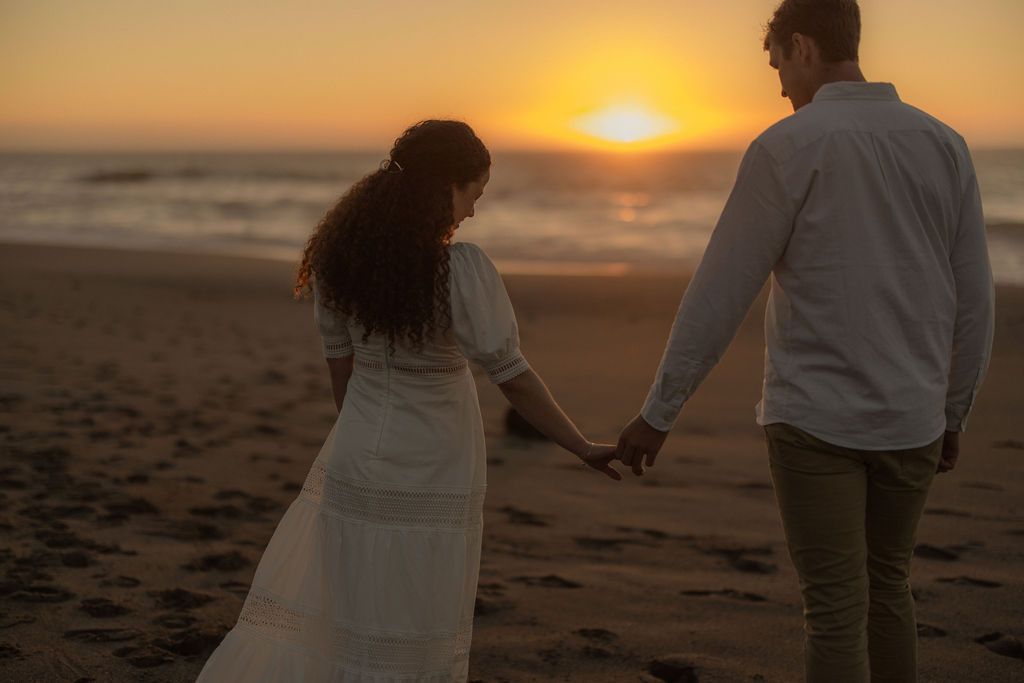 Couple holding hands and walking toward the sunset along the beach, romantic coastal engagement session