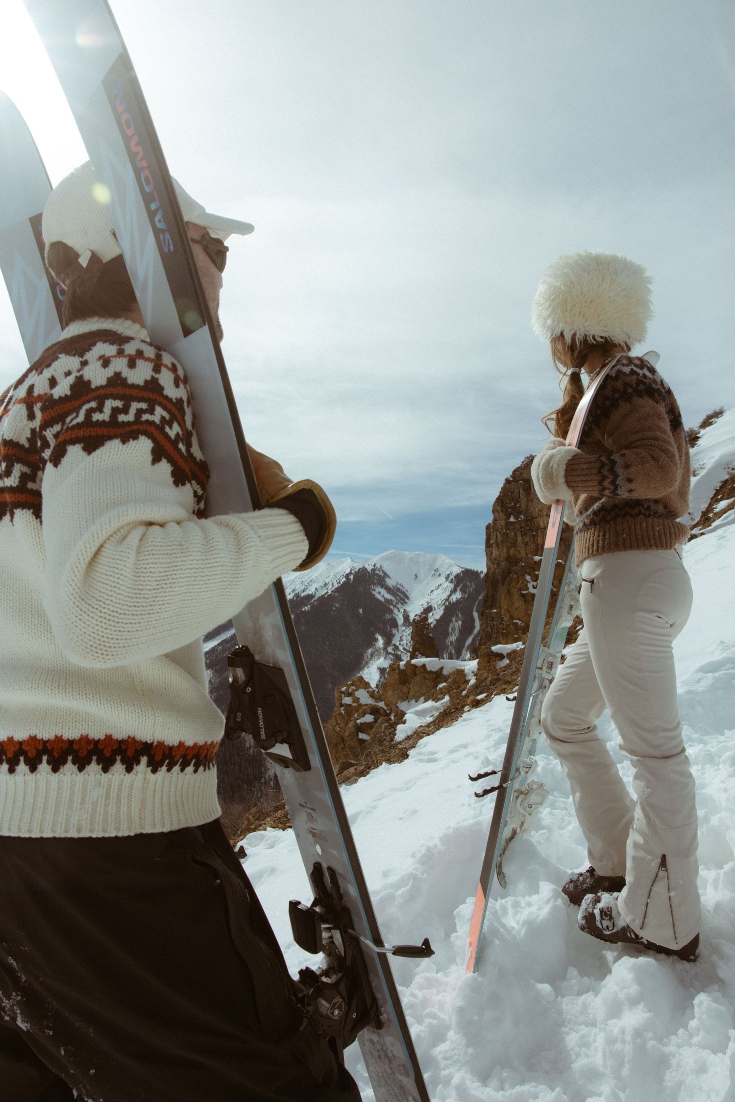 Couple in matching ski sweaters holding their skis on a snowy mountain slope, stylish retro winter engagement session