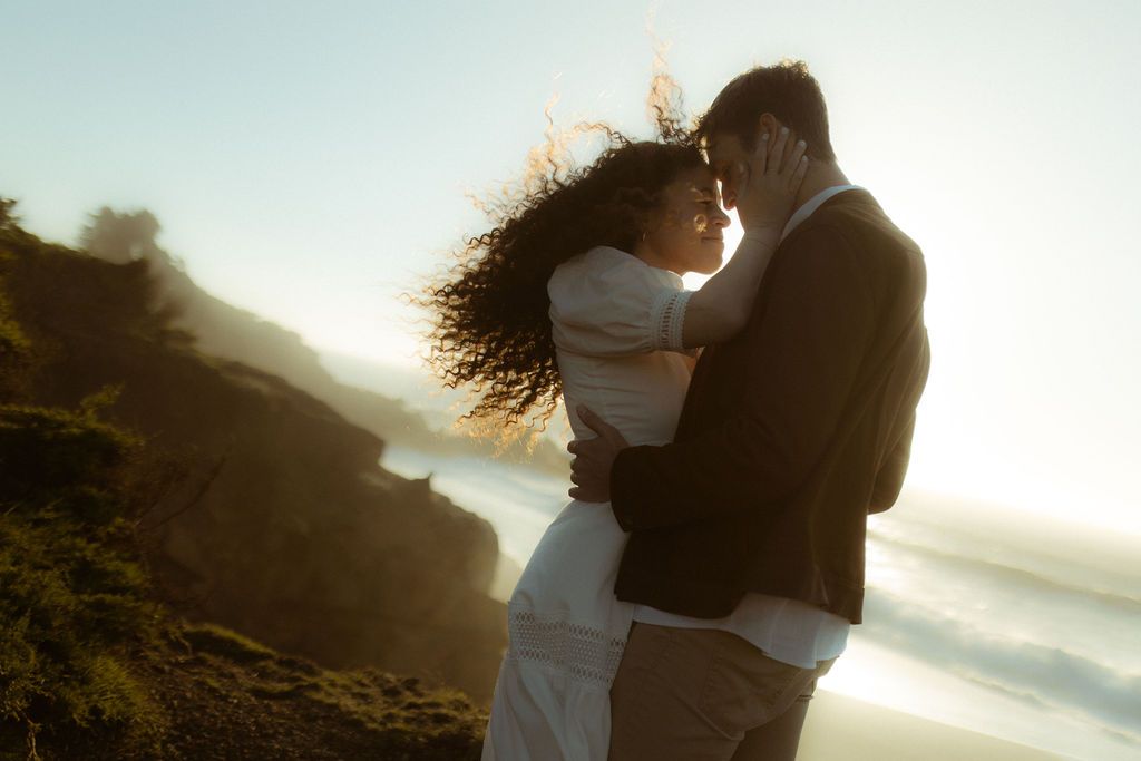 Couple resting forehead to forehead on a windswept coastal bluff at sunset, romantic elopement photography