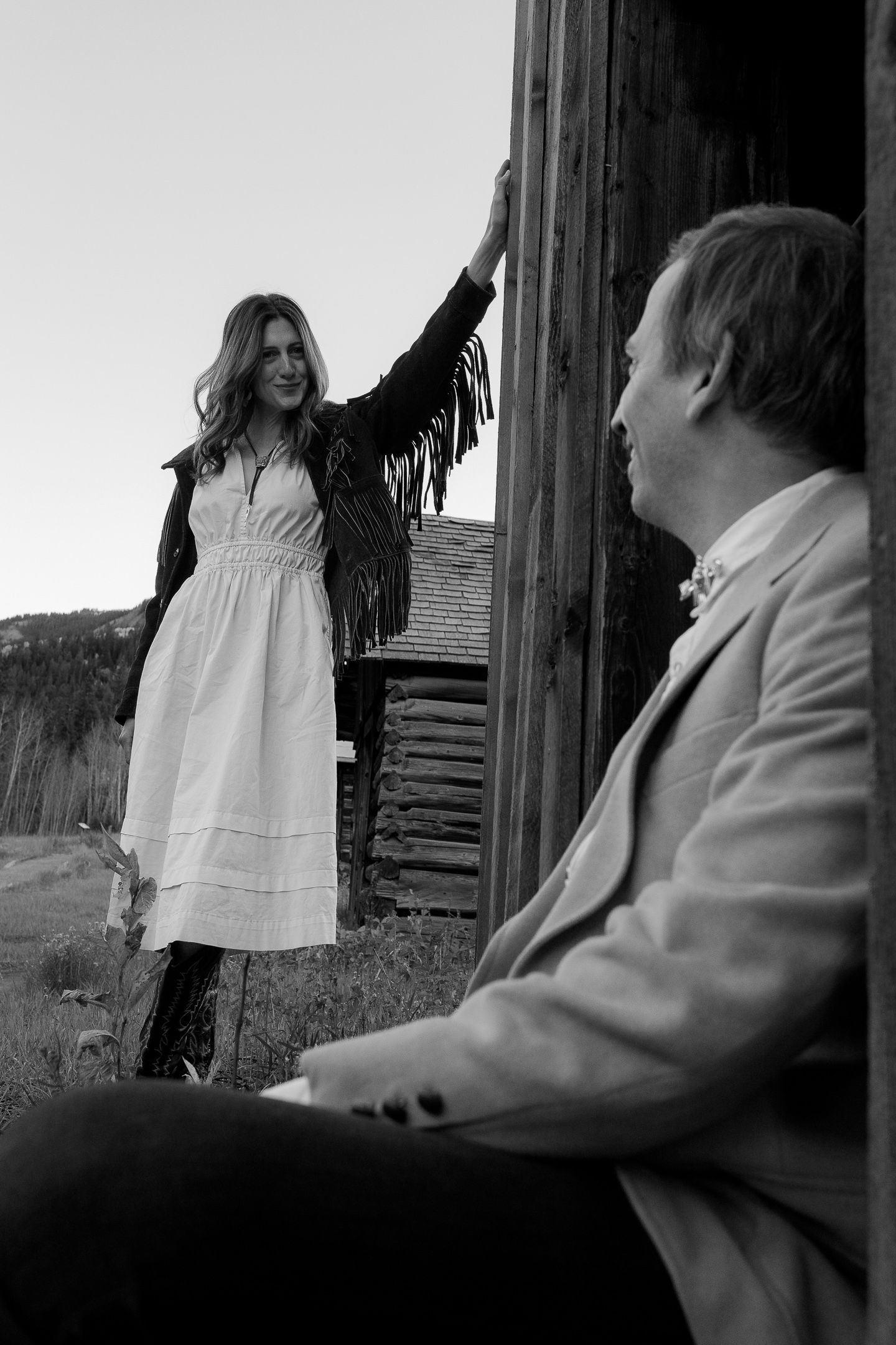 Black and white image of a couple by a weathered cabin, the woman in a white dress, timeless Mendocino engagement session