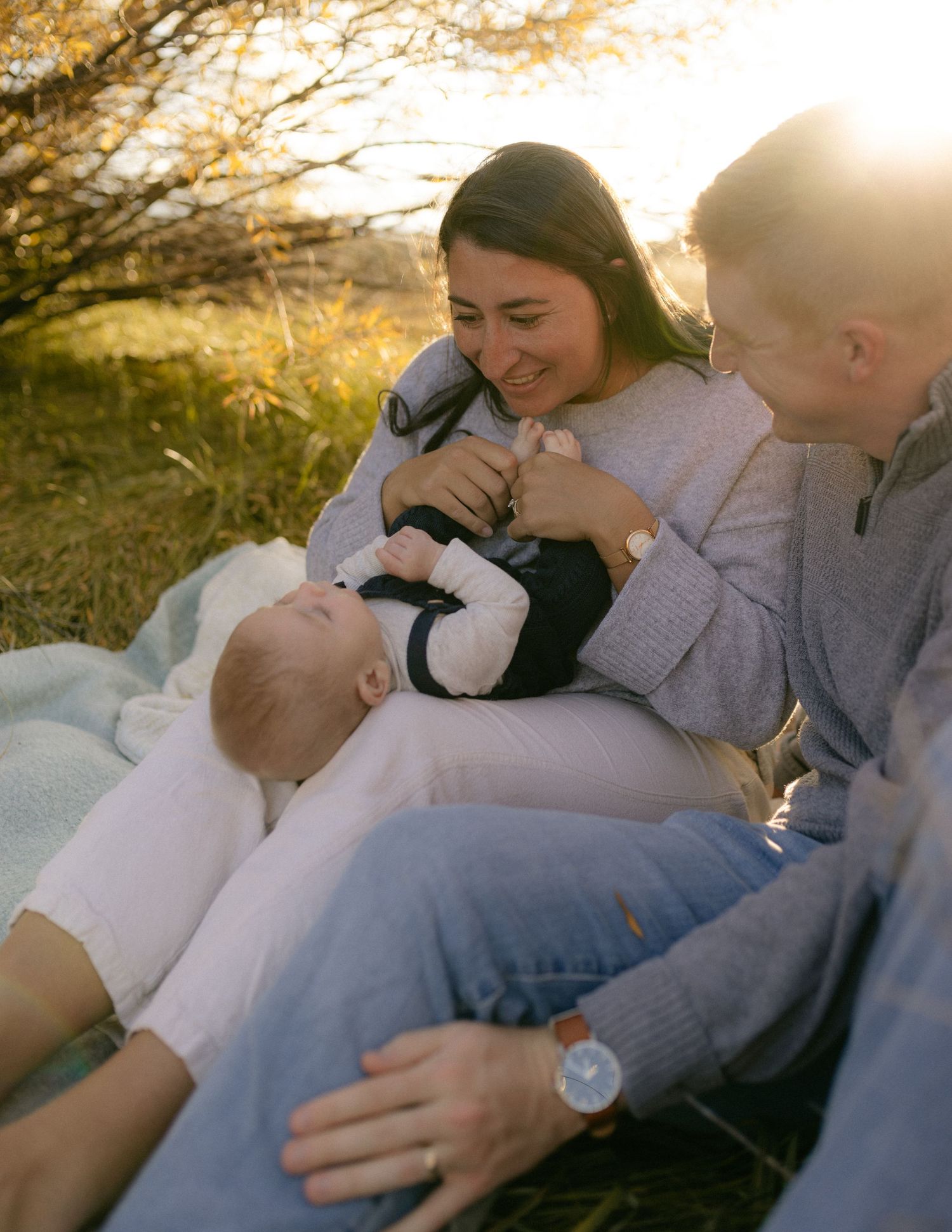 Parents cuddling their baby on a blanket in a golden autumn meadow at sunset, warm and tender family session