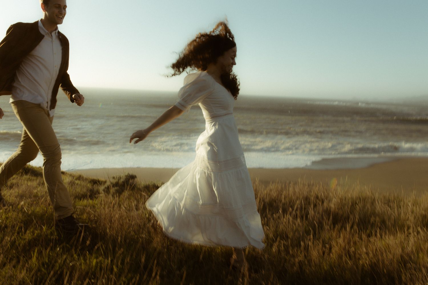 Couple playing on a coastal bluff at sunset, the bride's dress catching the wind, joyful engagement session