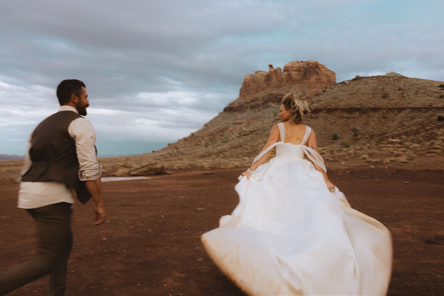 Groom and bride with billowing gown walking toward red rock buttes under a dramatic sky, adventurous desert elopement