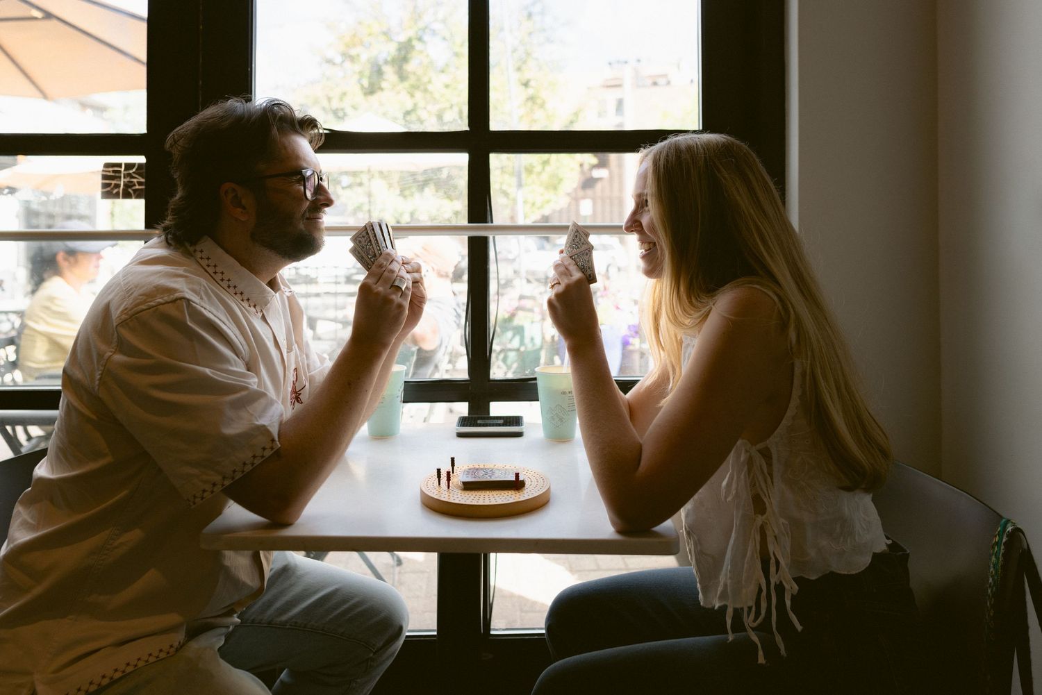 Couple sitting across a sunlit cafe table sharing a drink and laughing during a relaxed in-town engagement session