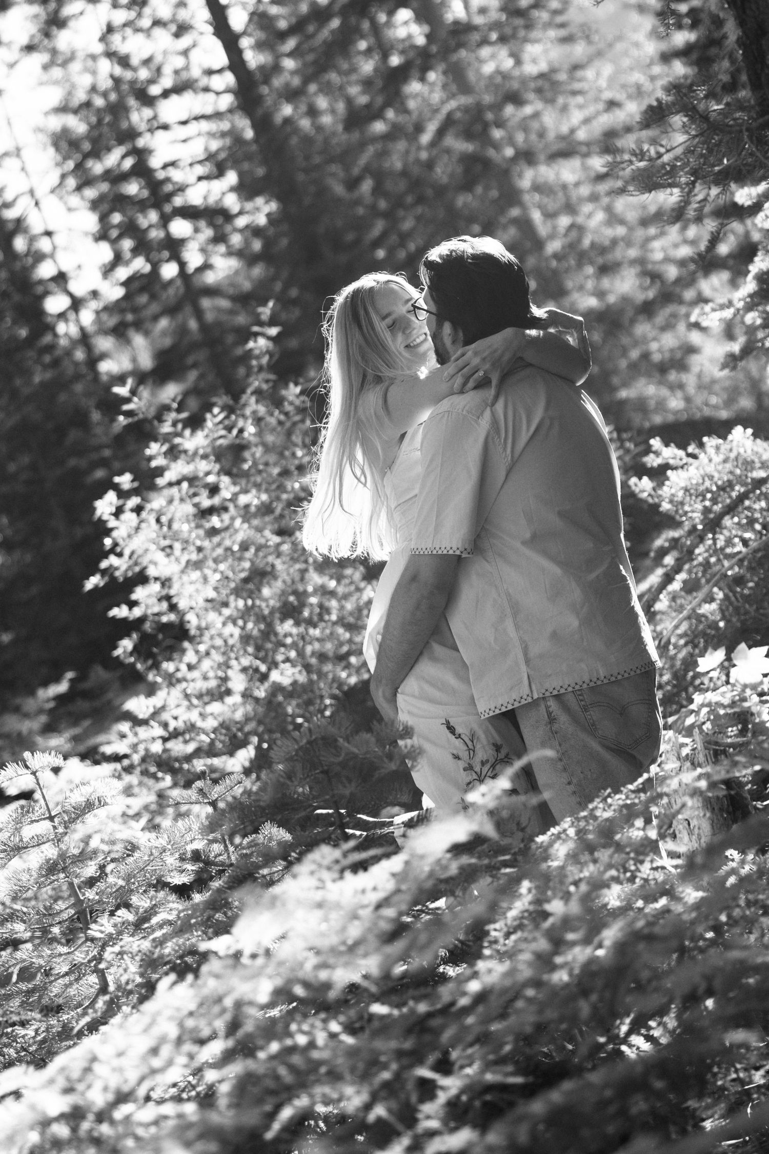 Black-and-white photo of a couple embracing among sunlit forest pines during a romantic mountain engagement session