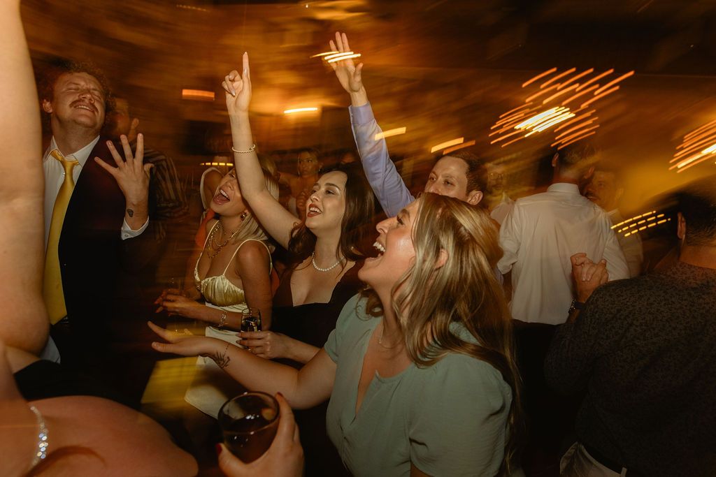 Guests dancing with hands raised on a warmly lit reception floor, joyful wedding celebration