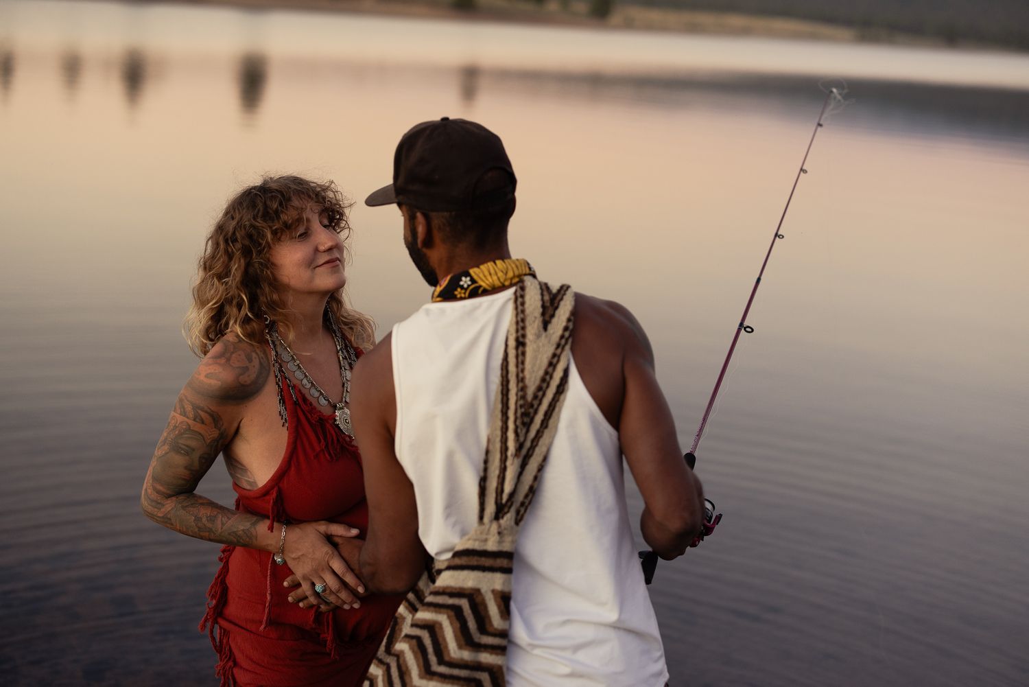 Couple embracing at the edge of a still lake at golden hour, one holding a fishing rod, laid-back engagement session