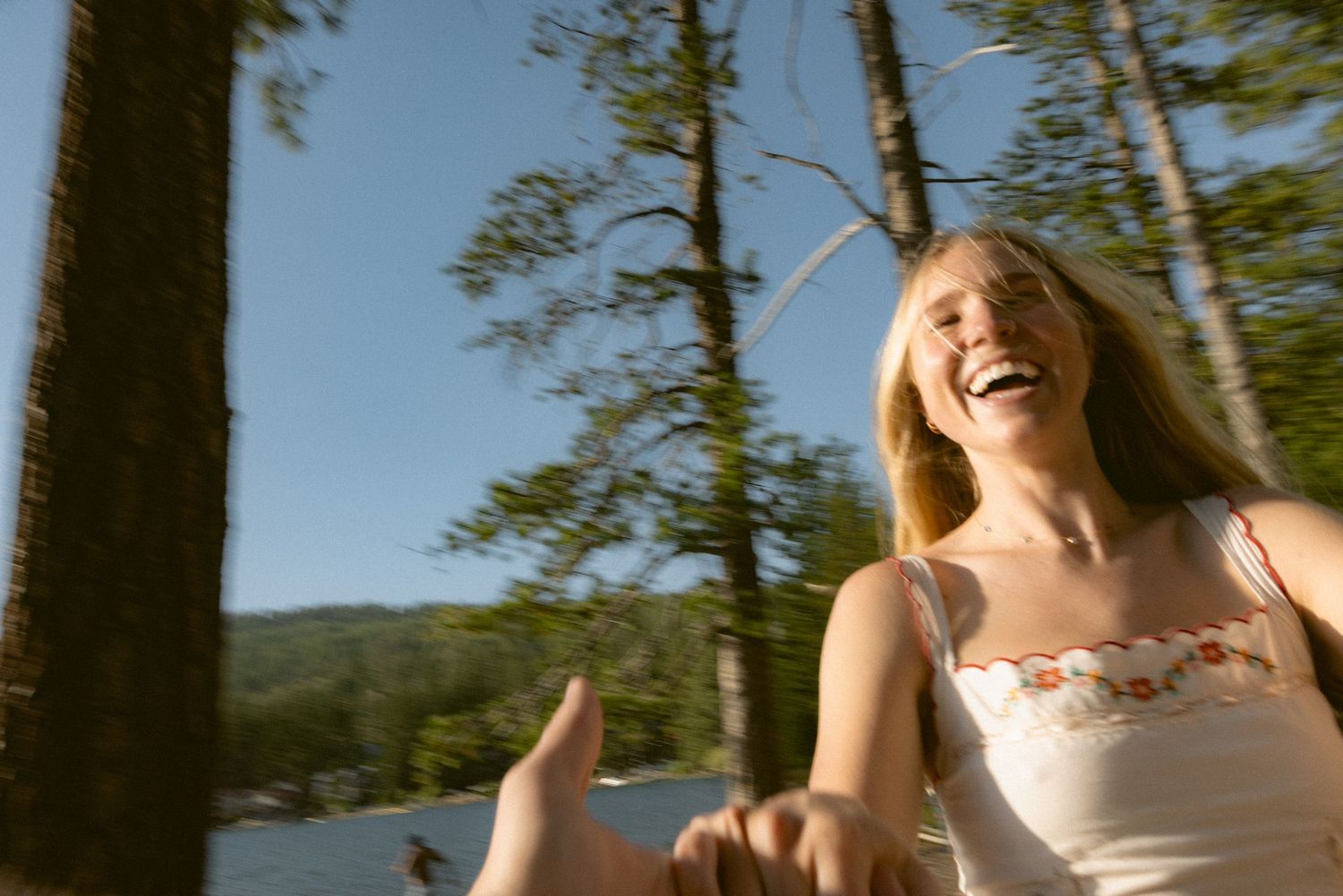 Laughing woman in a white sundress led by the hand through a sunlit pine forest beside a lake, joyful engagement story session